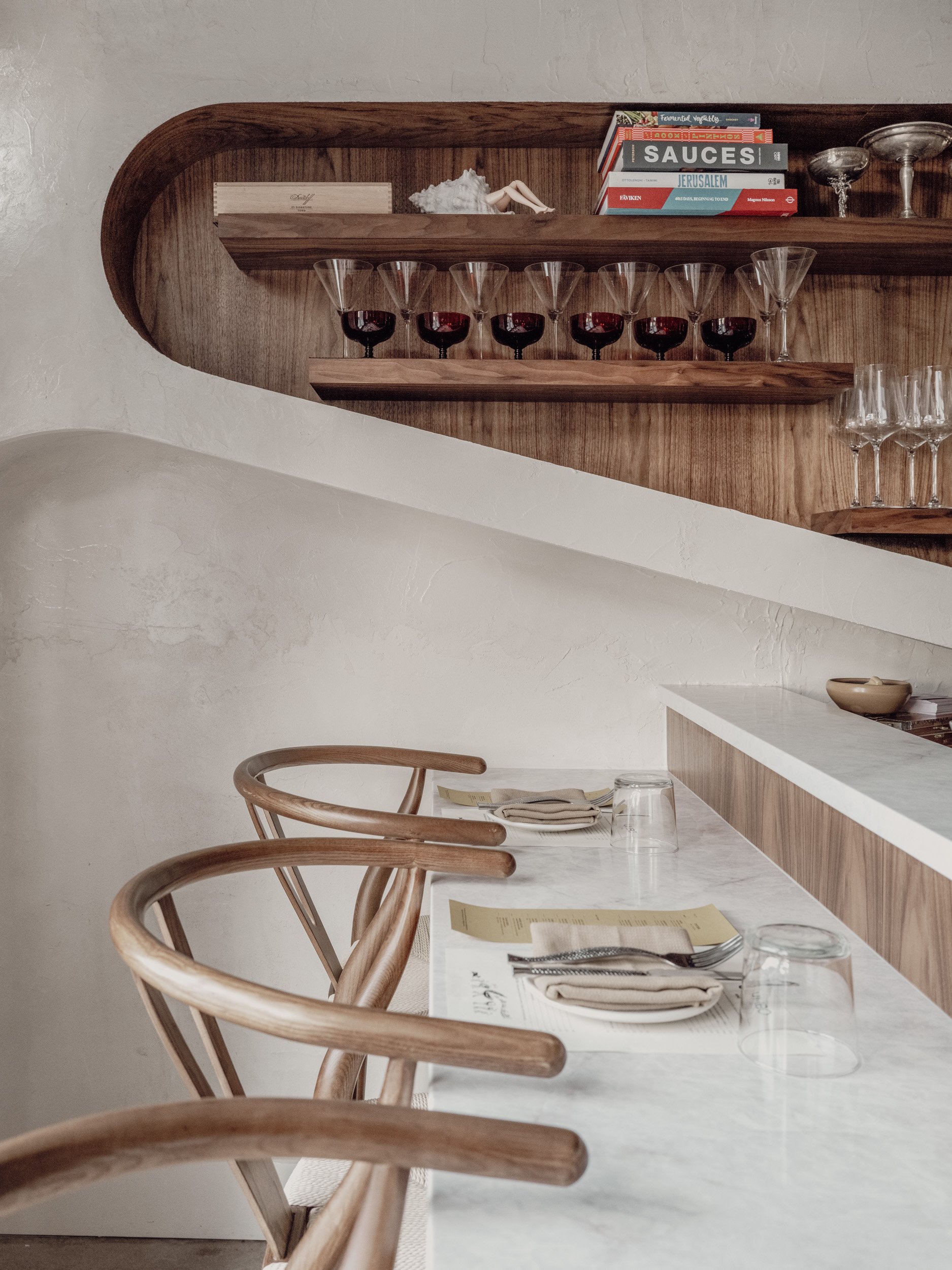 Interior of restaurant dining space with counter stools, warm woods, and soft ceiling textures.