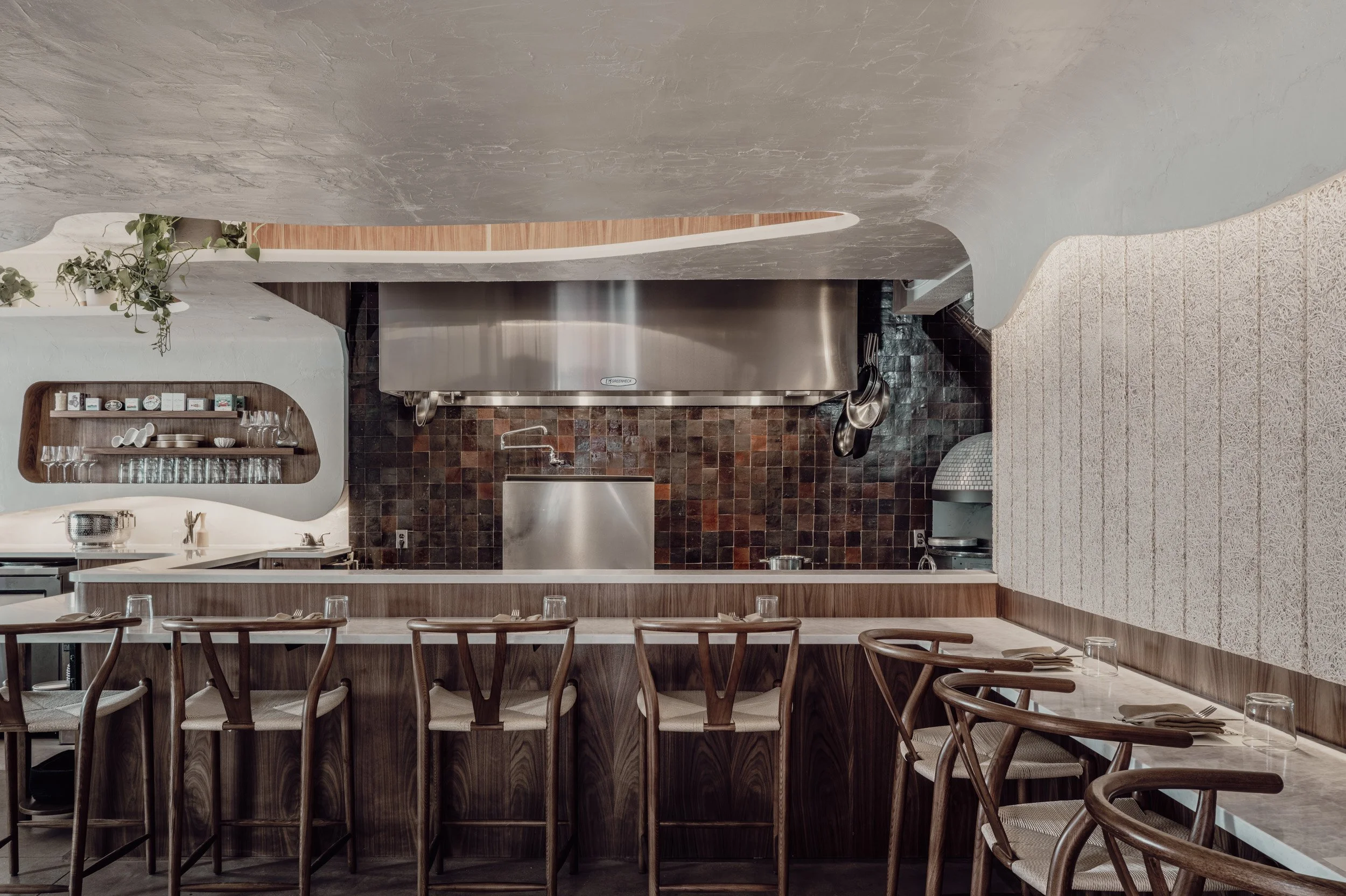 Interior of restaurant dining space with counter stools, warm woods, and soft ceiling textures.