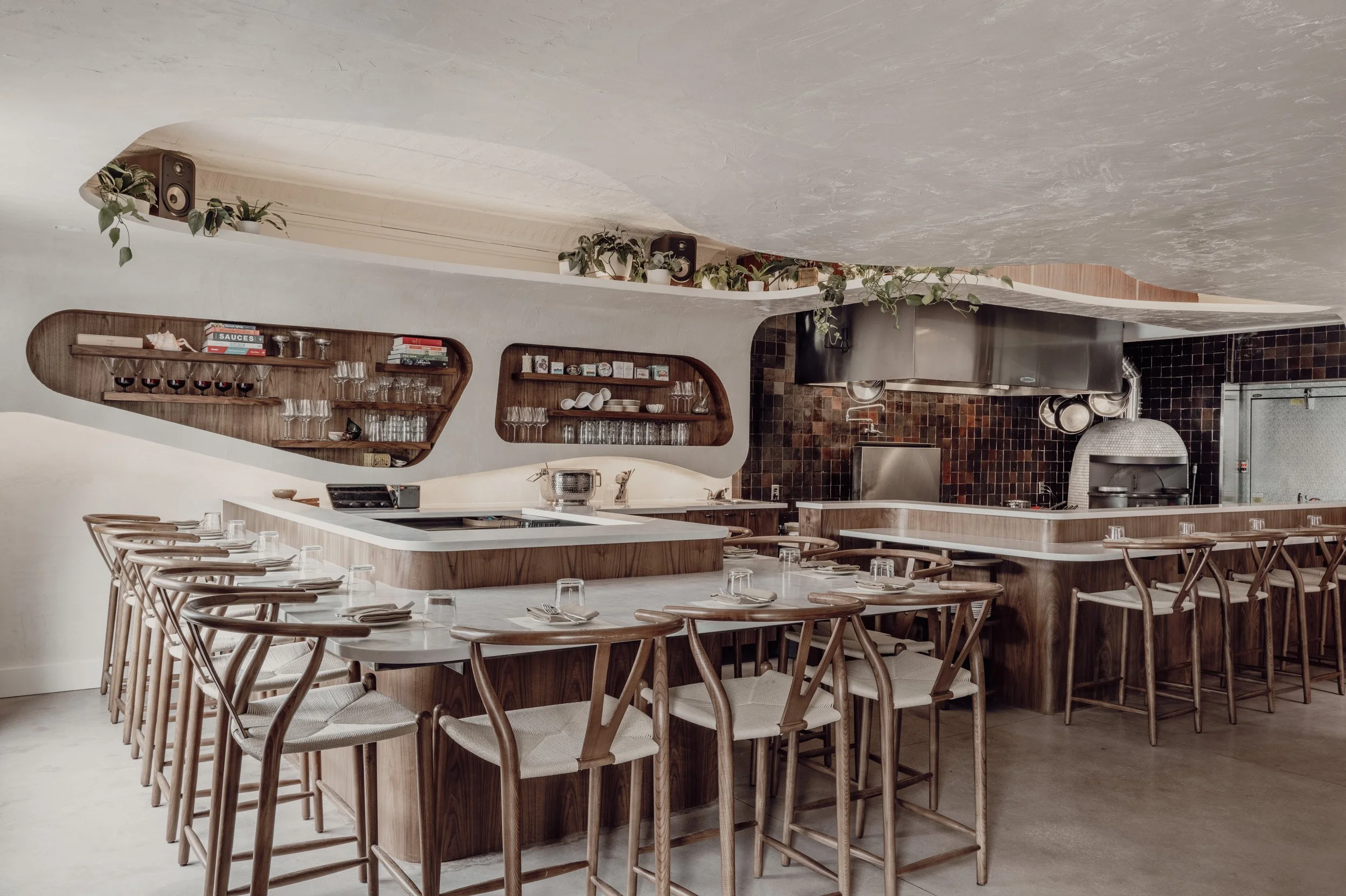 Interior of restaurant dining space with counter stools, warm woods, and soft ceiling textures.