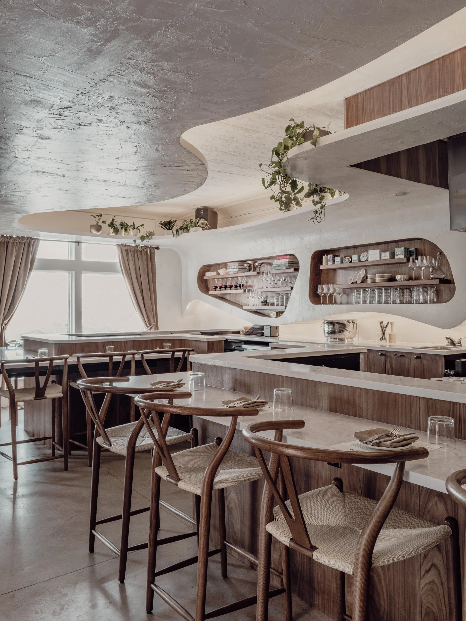 Interior of restaurant dining space with counter stools, warm woods, and soft ceiling textures.
