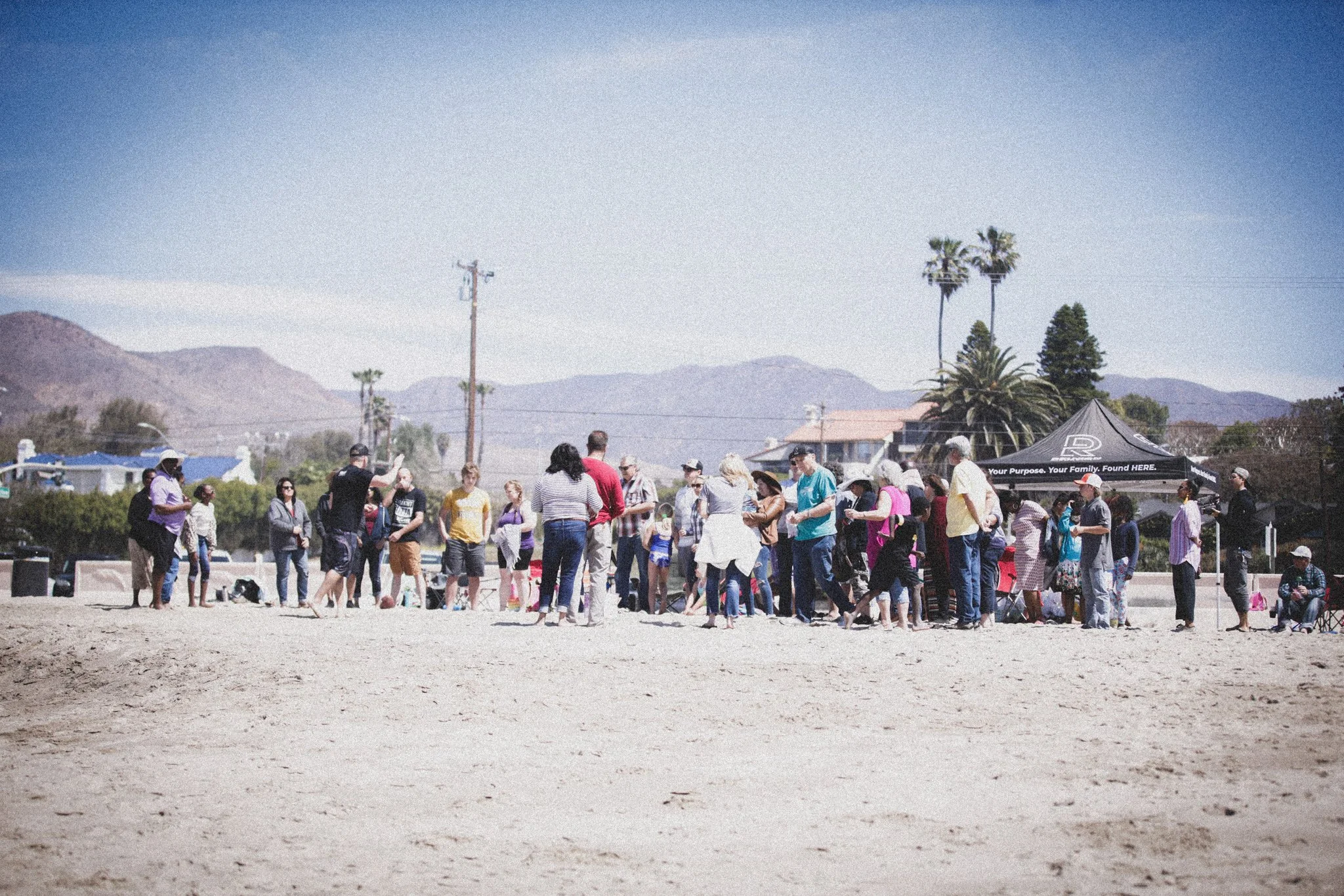 Group of people gathered outdoors under a black tent with a slogan, with mountains and palm trees in the background on a sunny day.