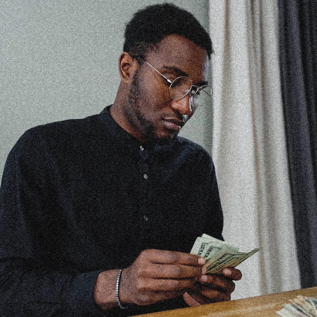 A young man with glasses and a black shirt counting dollar bills at a table.