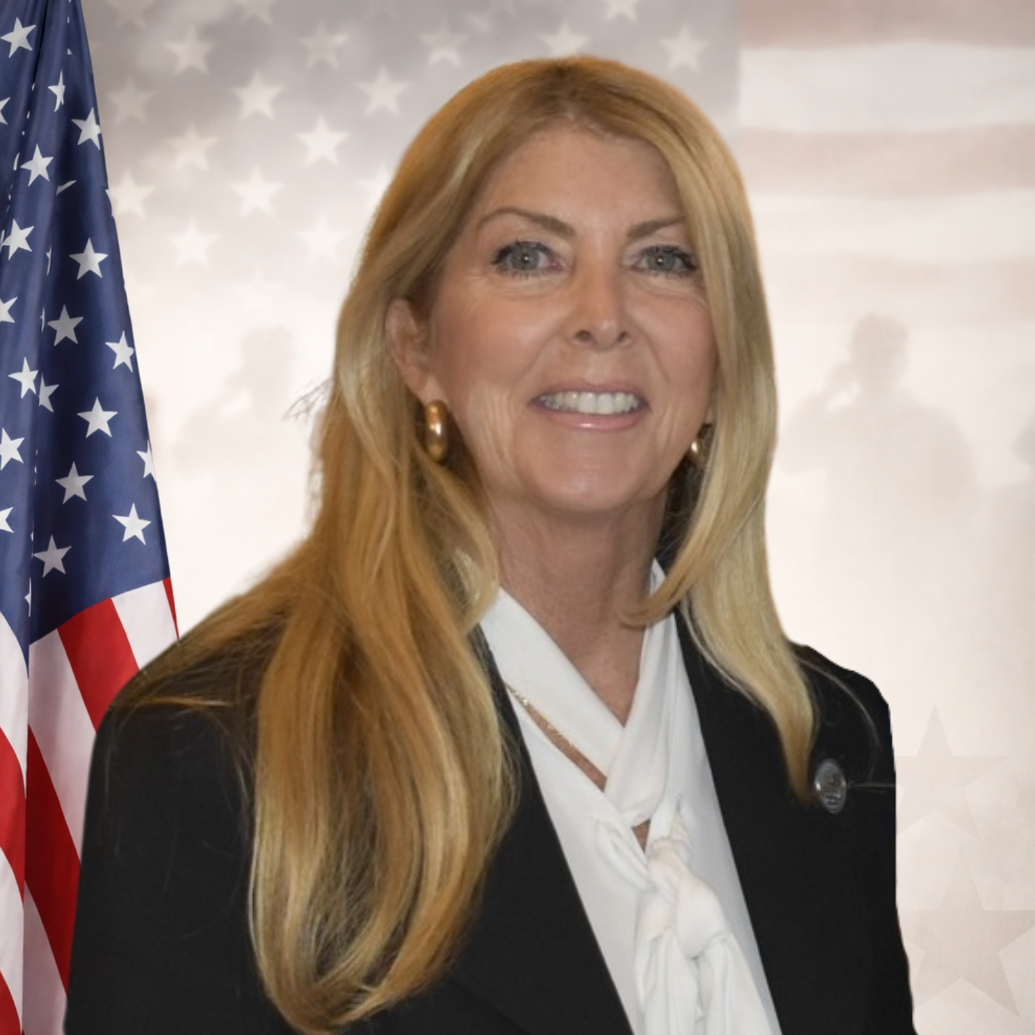 A smiling middle-aged woman with long blonde hair, wearing a black blazer, white blouse, and gold earrings, at a professional event.