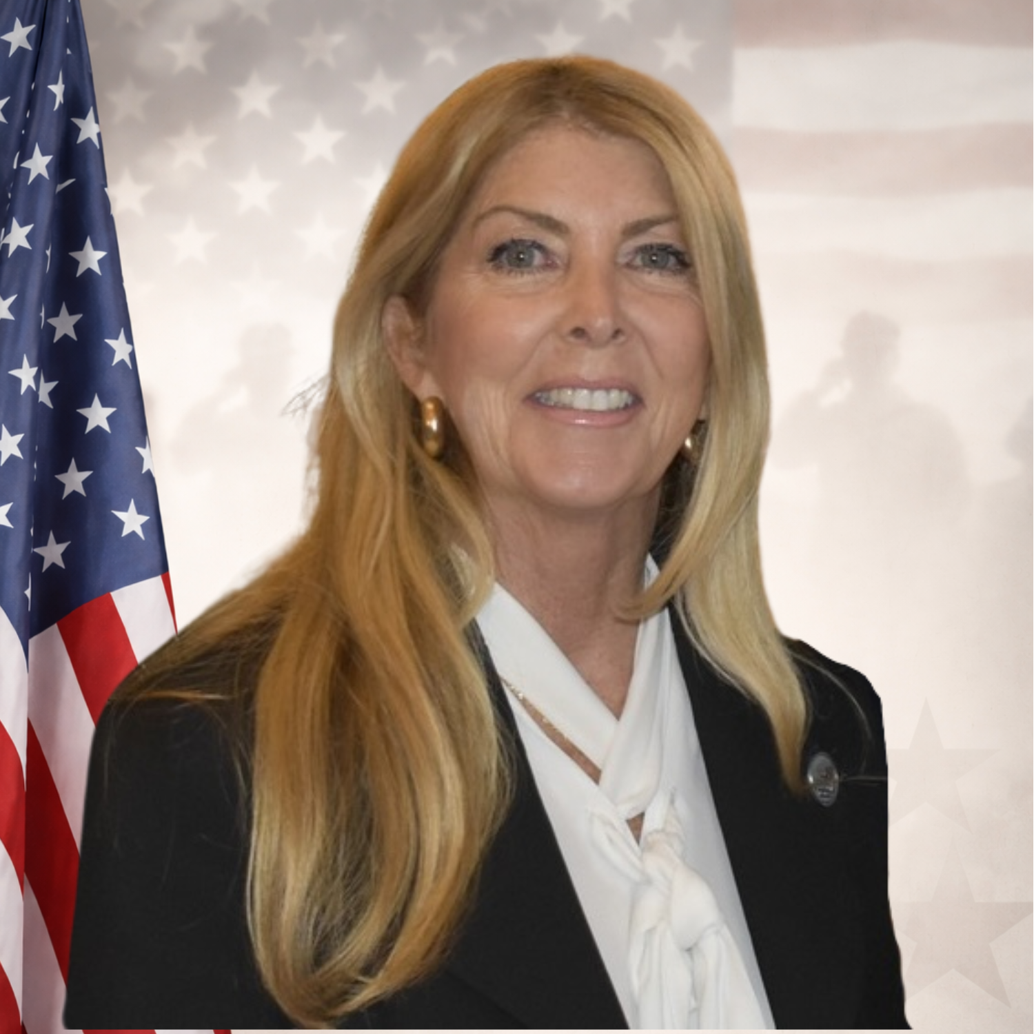 A smiling middle-aged woman with long blonde hair, wearing a black blazer, white blouse, and gold earrings, at a professional event.