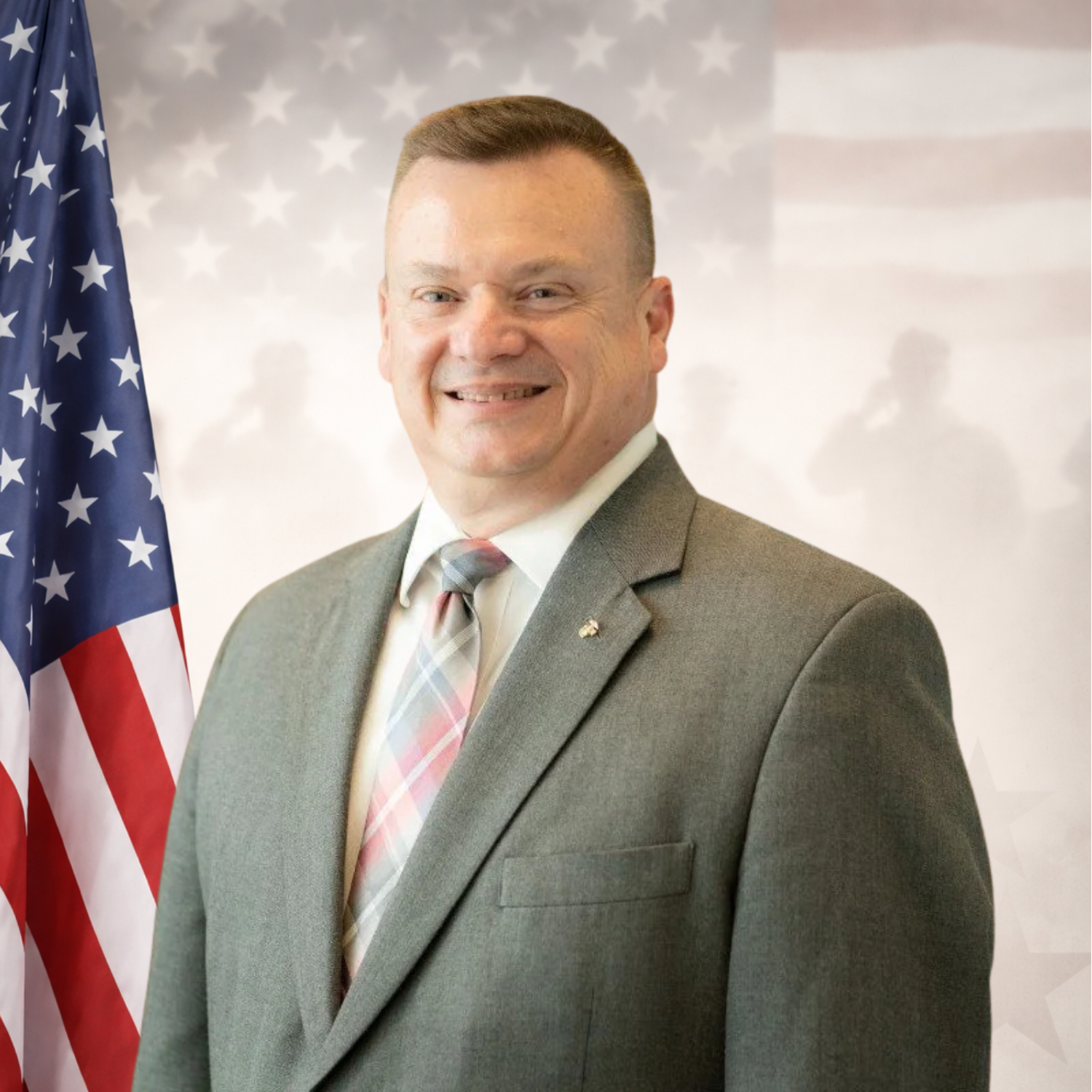 A man with glasses and a white beard wearing a black polo shirt with a company logo standing in front of a blue wall.