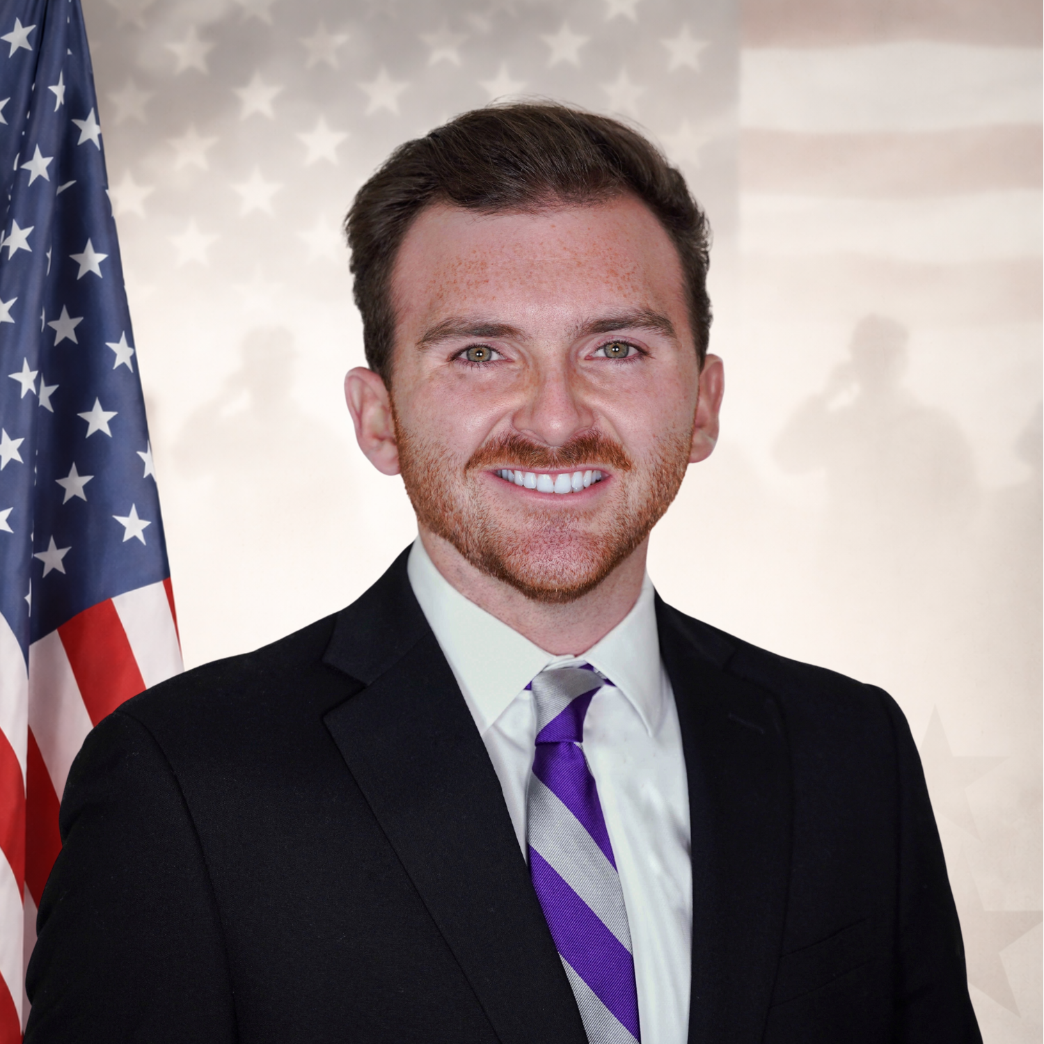 A smiling man wearing glasses, a blue suit, and a blue tie, standing in front of a window with trees outside.