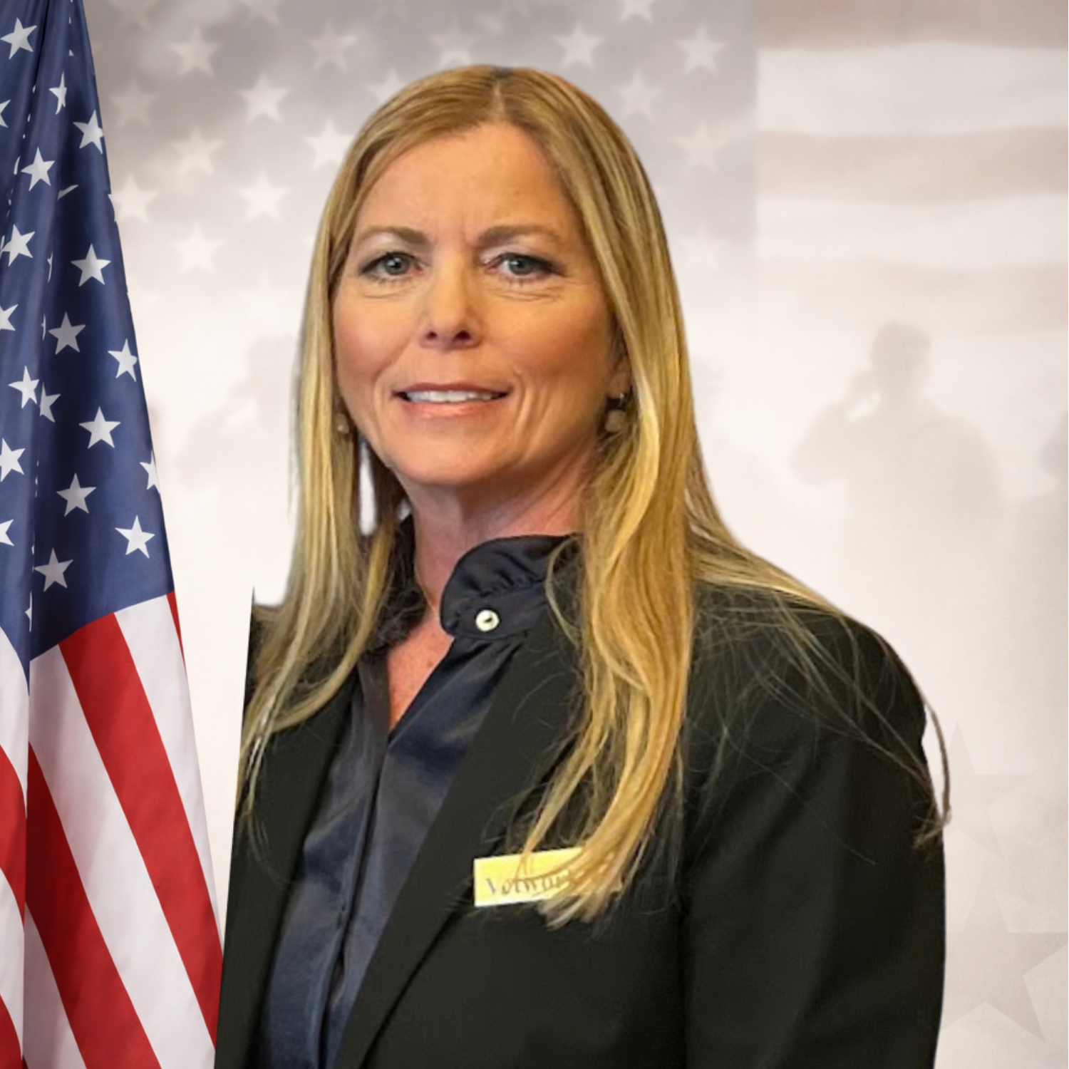 A middle-aged woman with long blonde hair, wearing a black blazer and a dark blouse, standing against a blue background.