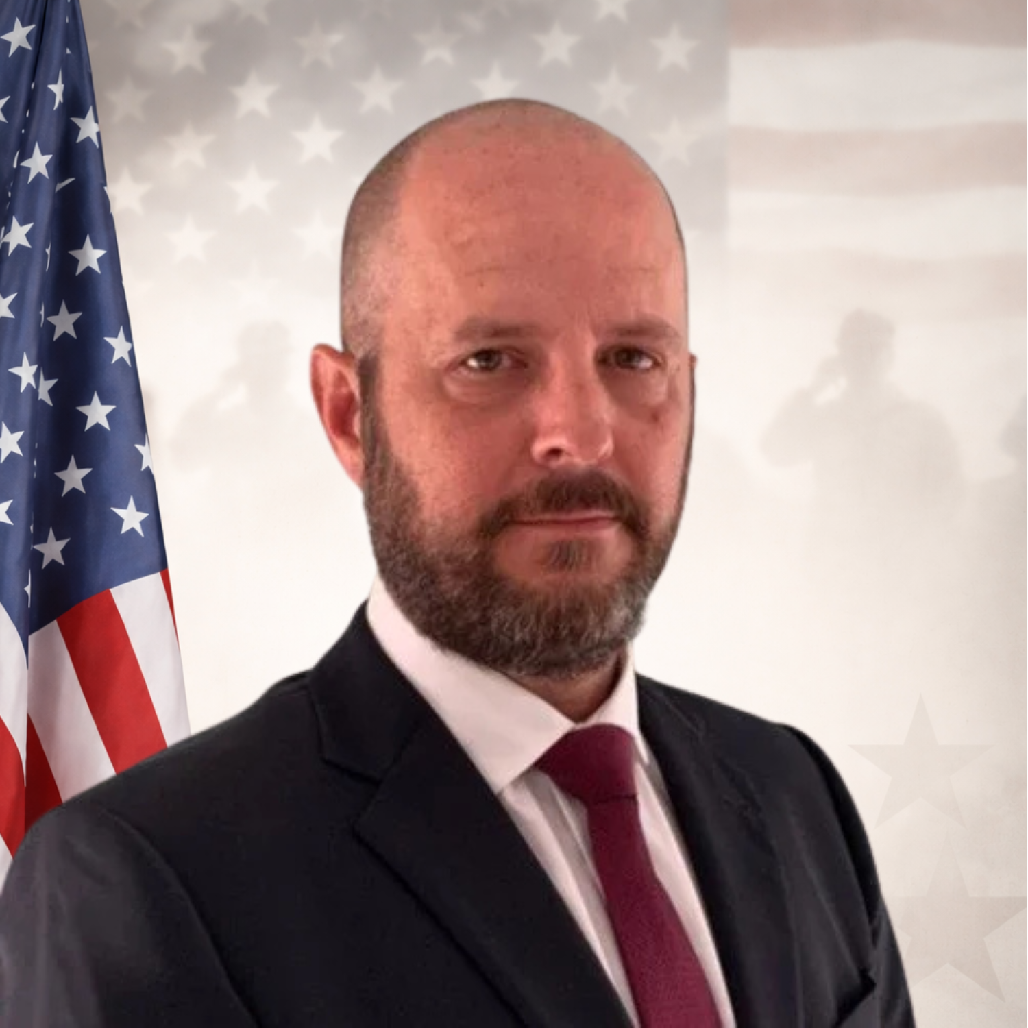 A man with a beard and bald head wearing a black suit with a white shirt and a maroon tie, standing against a plain light-colored background.