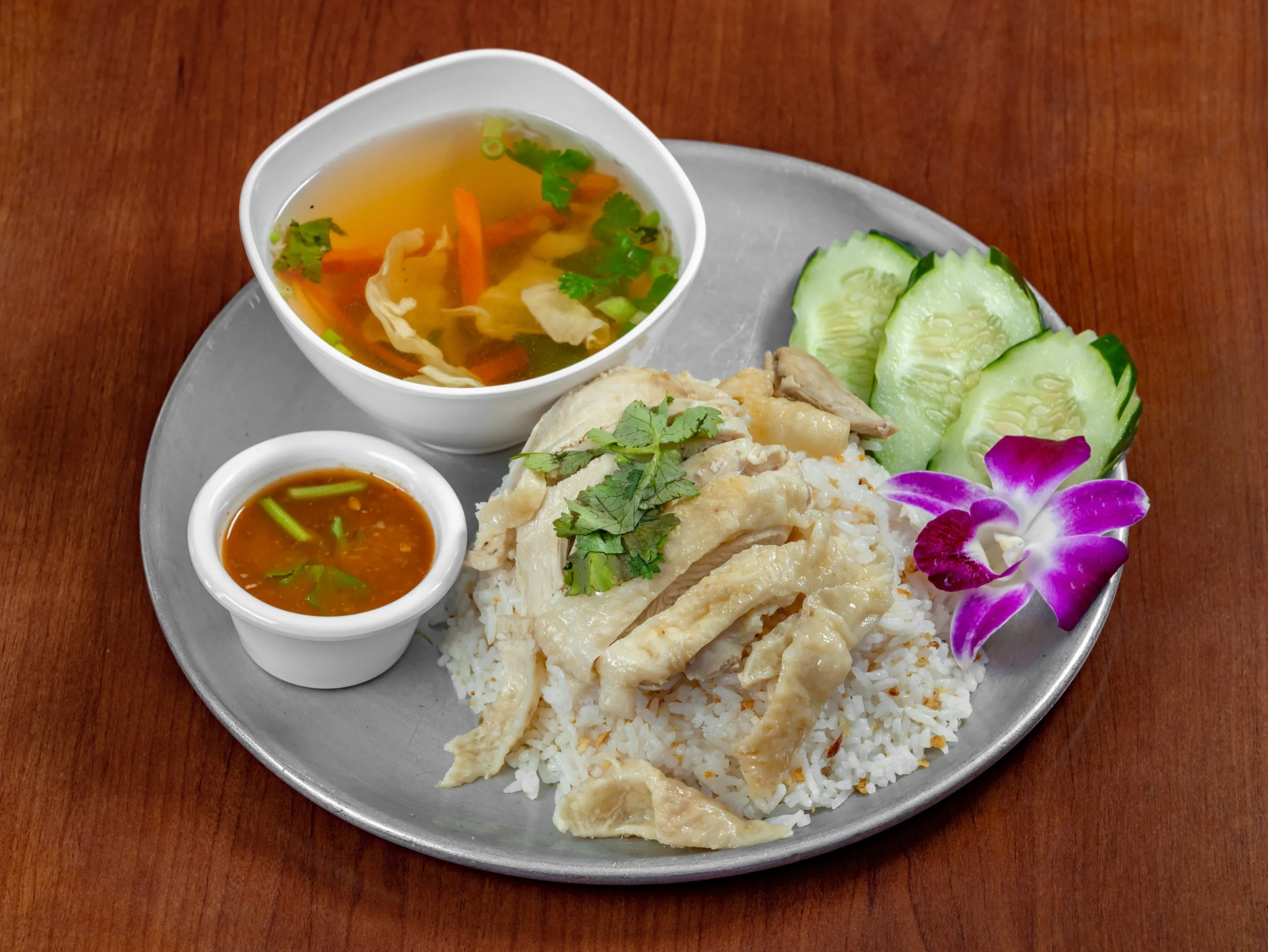 Khao Mun Gai, Thai-style steamed chicken served over fragrant seasoned jasmine rice, accompanied by a savory ginger-soy dipping sauce, fresh cucumber, cilantro, and a light chicken broth.