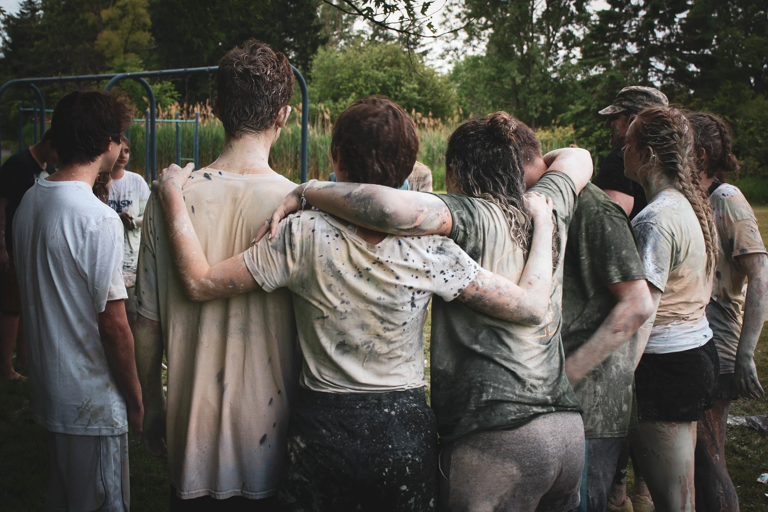 New Testimonials; group of teenagers covered in mud and paint, in a circle with their arms around each other