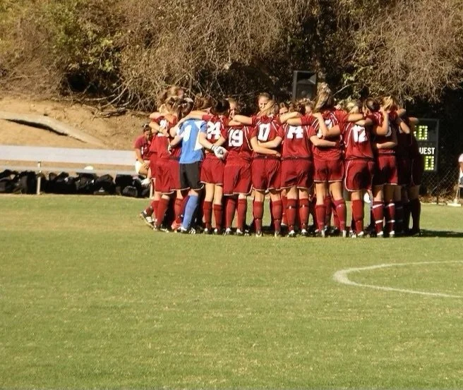 Group Services, Specialized Services; Group of female soccer players in a huddle with arms wrapped around one another in red uniforms on a soccer field