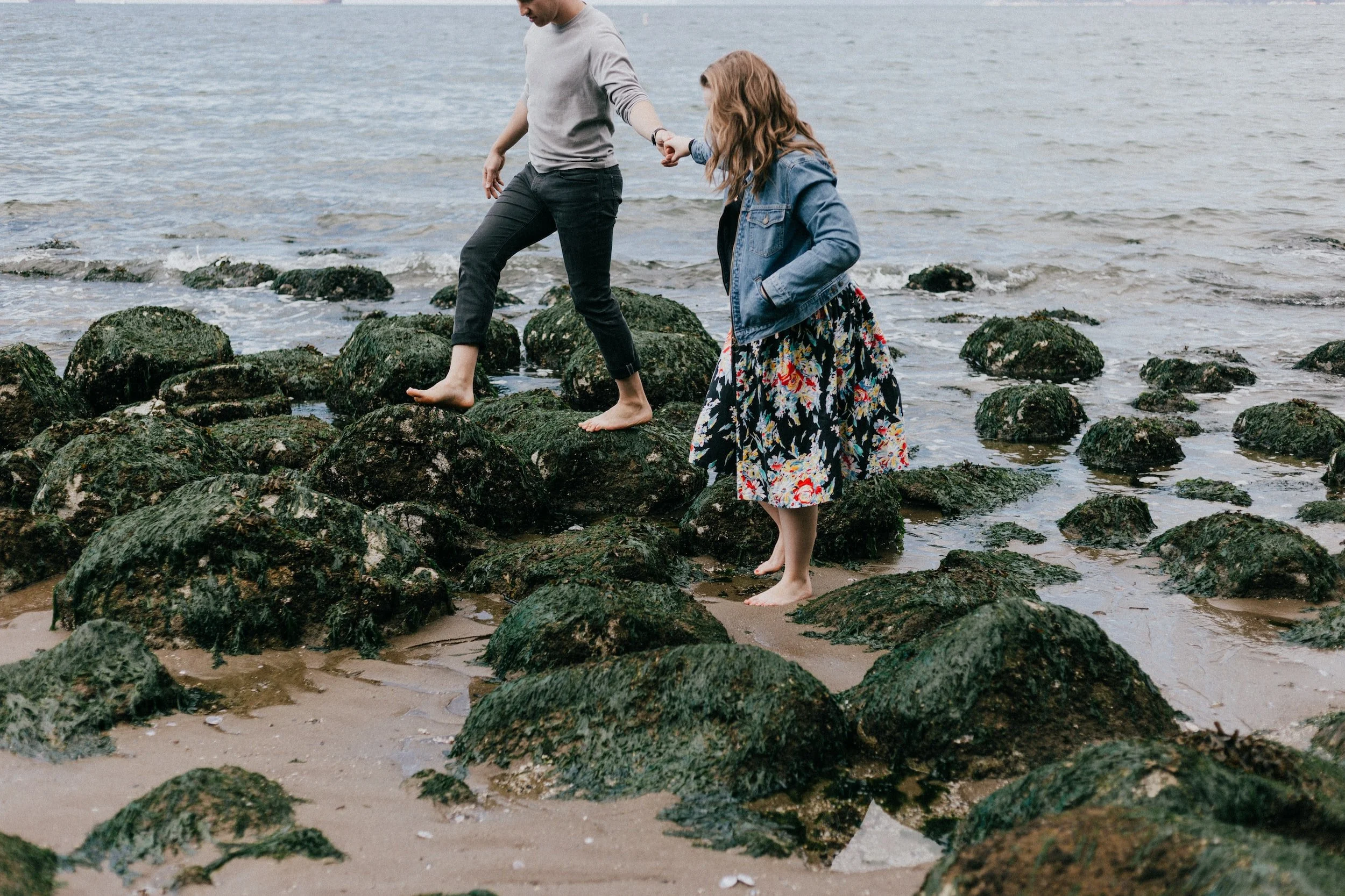 Two people walking barefoot on a rocky, seaweed-covered beach.