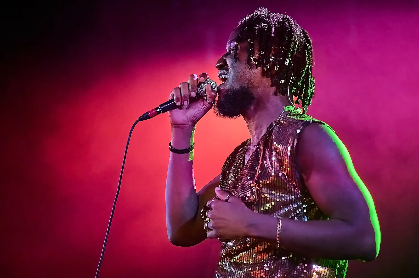 Singer performing on stage with a microphone, wearing a glittery top, against a colorful background.