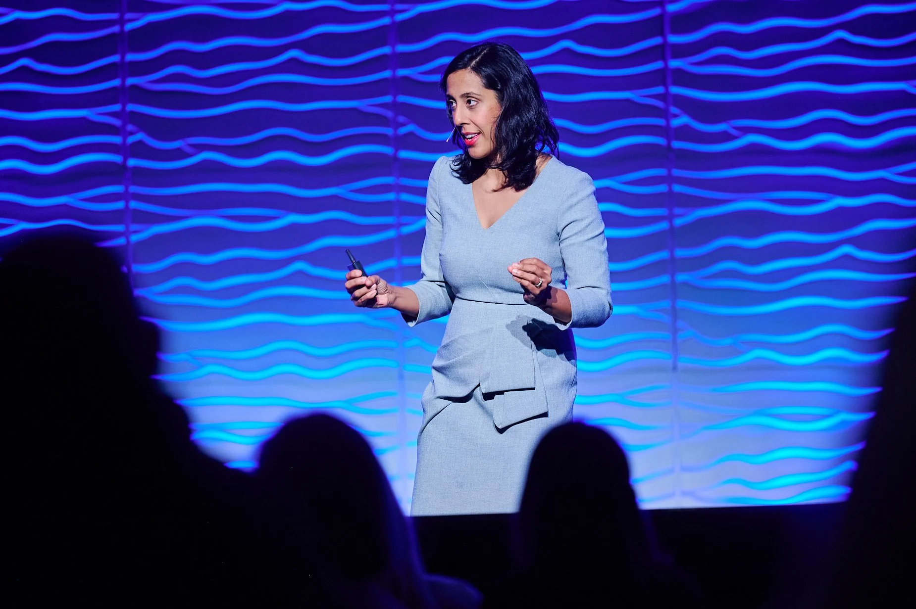 Woman giving a presentation on stage with a microphone, against a wavy blue background, holding a remote clicker.