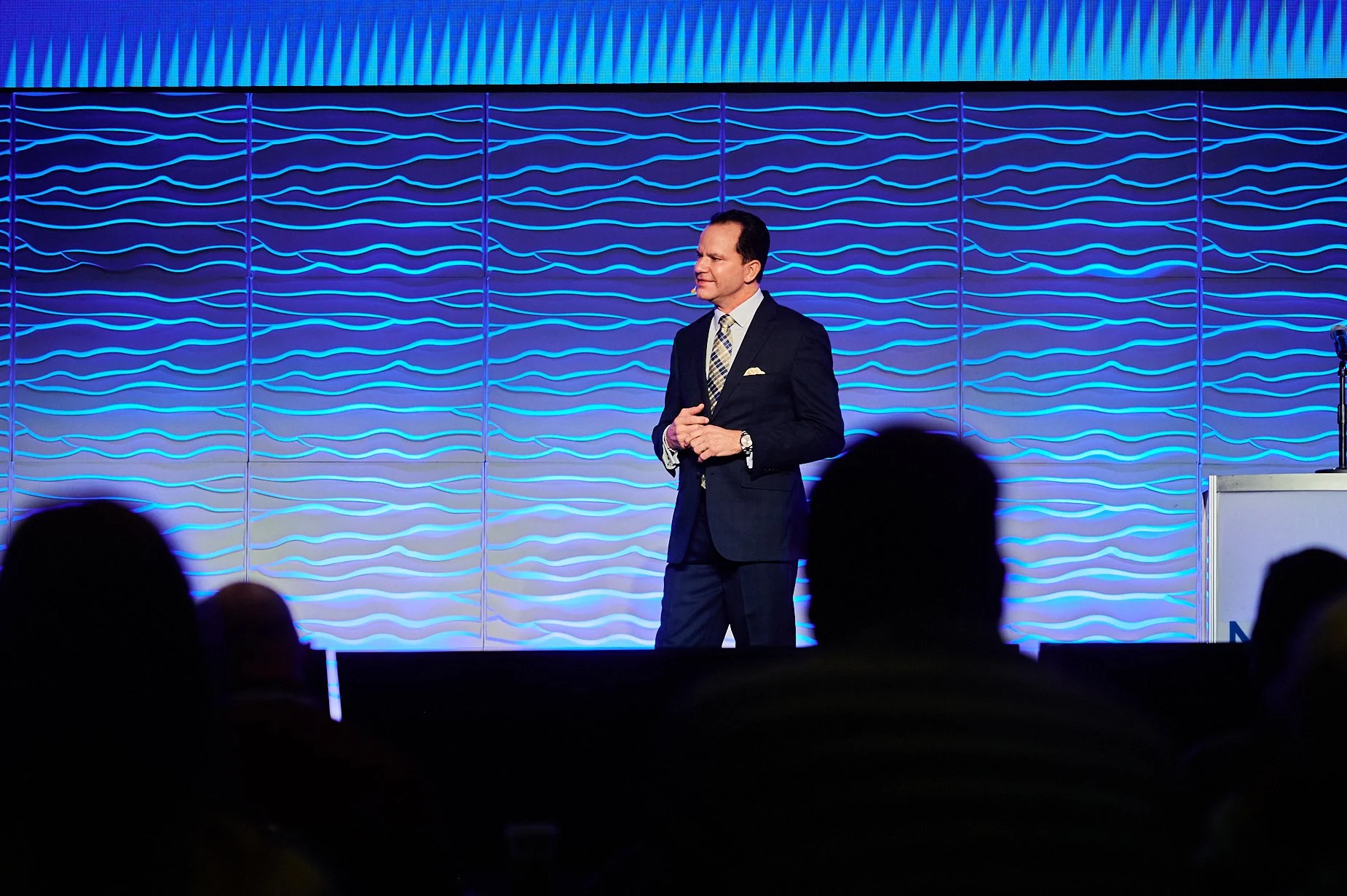 Man in suit speaking on stage with blue wave-patterned backdrop