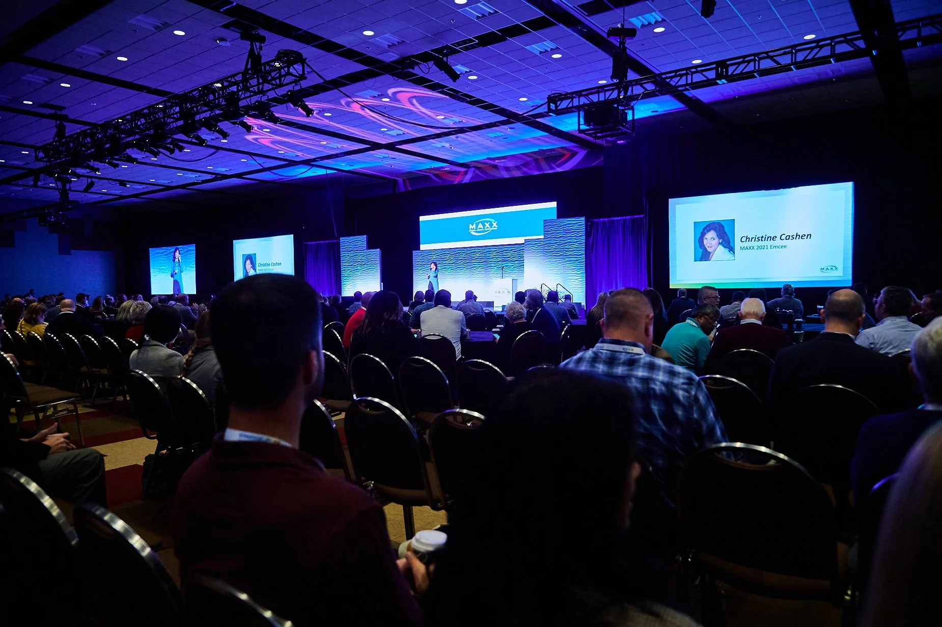 Conference audience listening to a speaker on stage with presentation screens in a large hall.