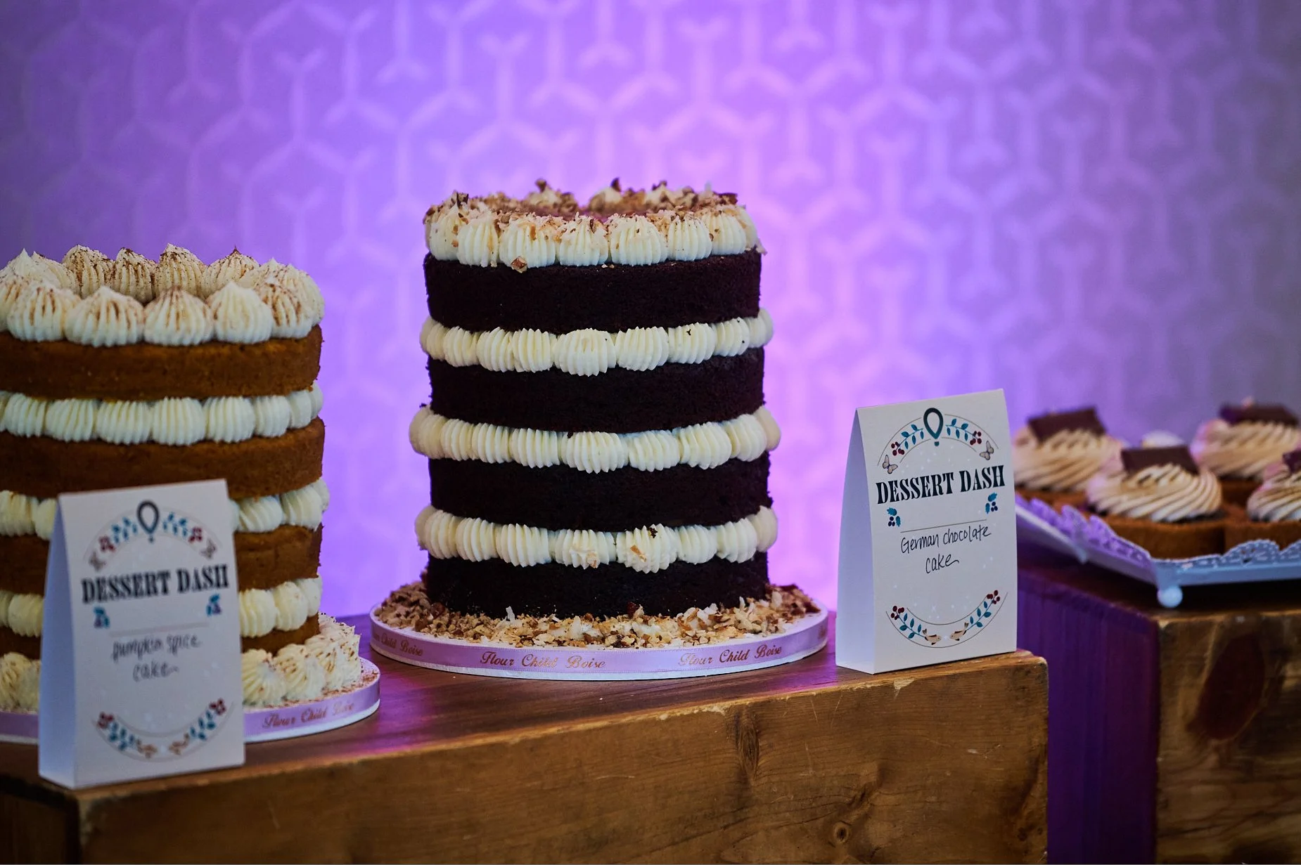 Display of tiered cakes with vanilla frosting, labeled as pumpkin spice and German chocolate cakes, on wooden table.