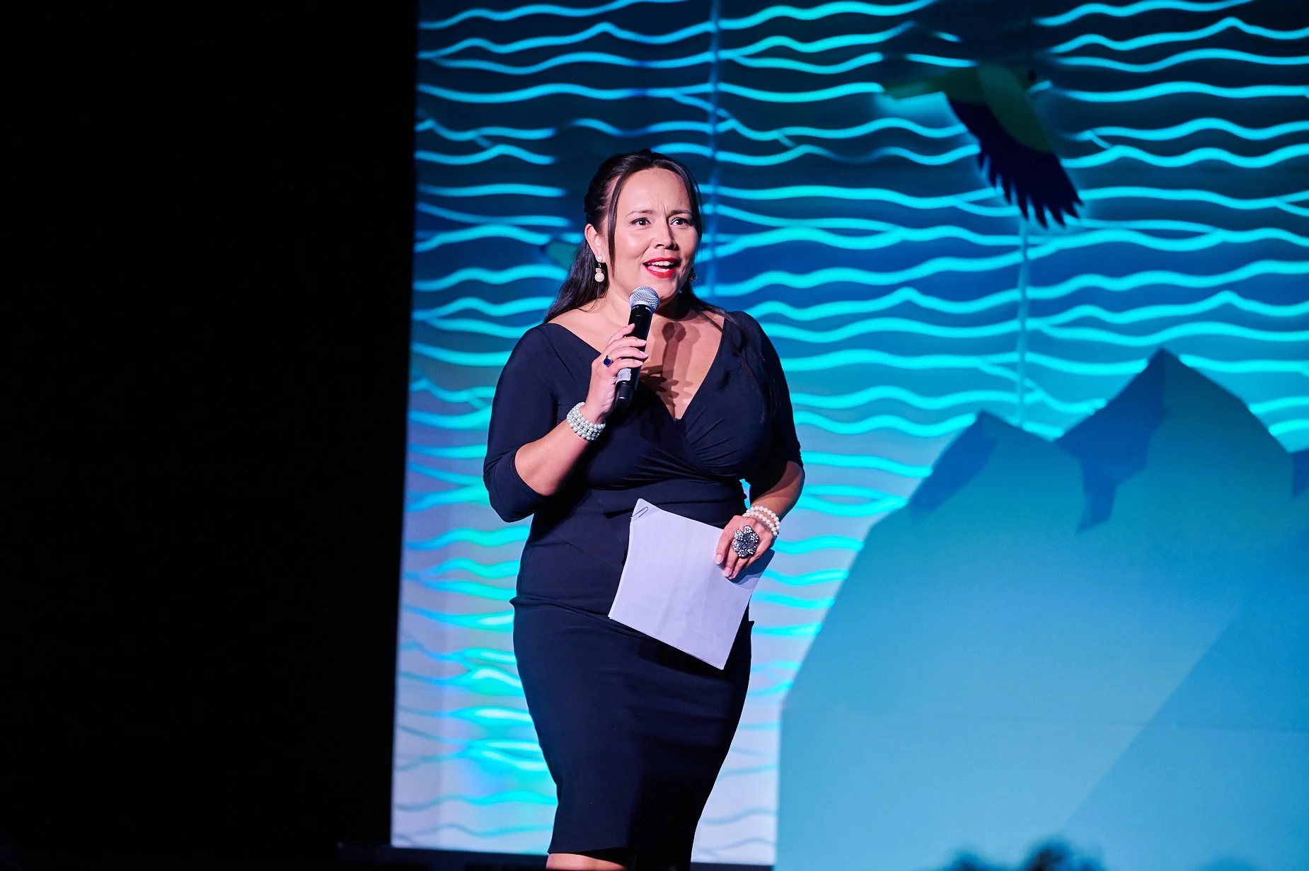 Woman in black dress speaking on stage with microphone, holding paper, against a blue wave-patterned backdrop.