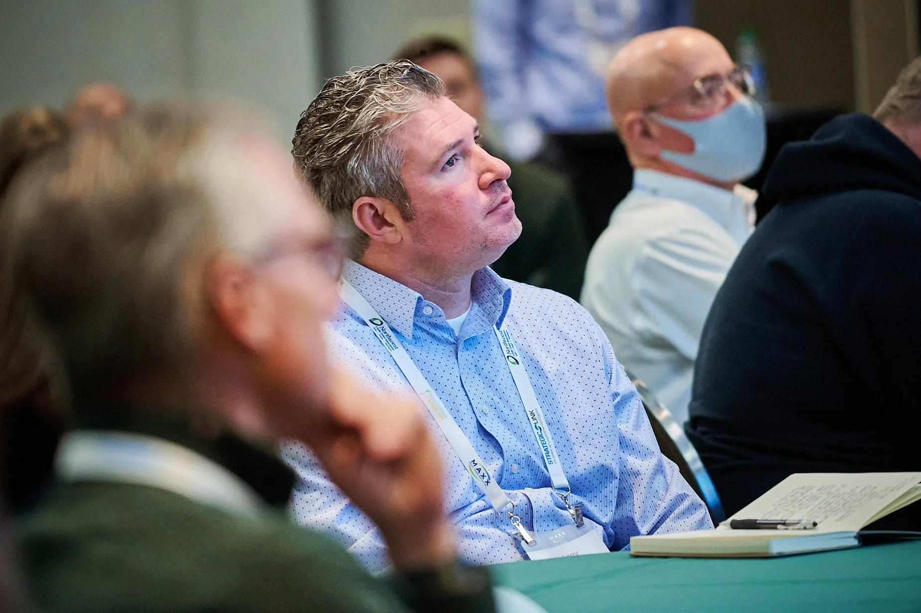 People at a conference listening attentively, wearing lanyards with name tags, and one person wearing a face mask.