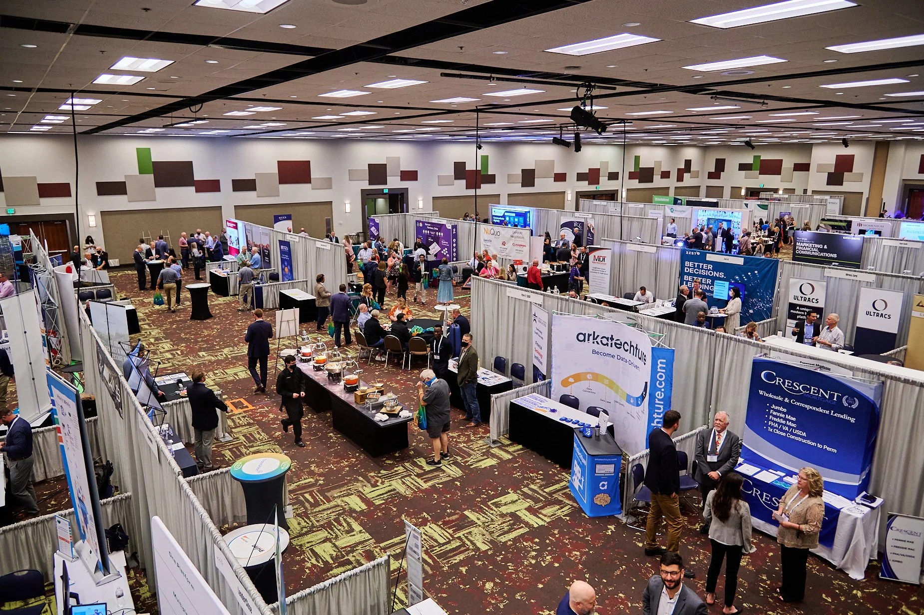 A busy trade show with various booths and attendees interacting, set in a large hall.