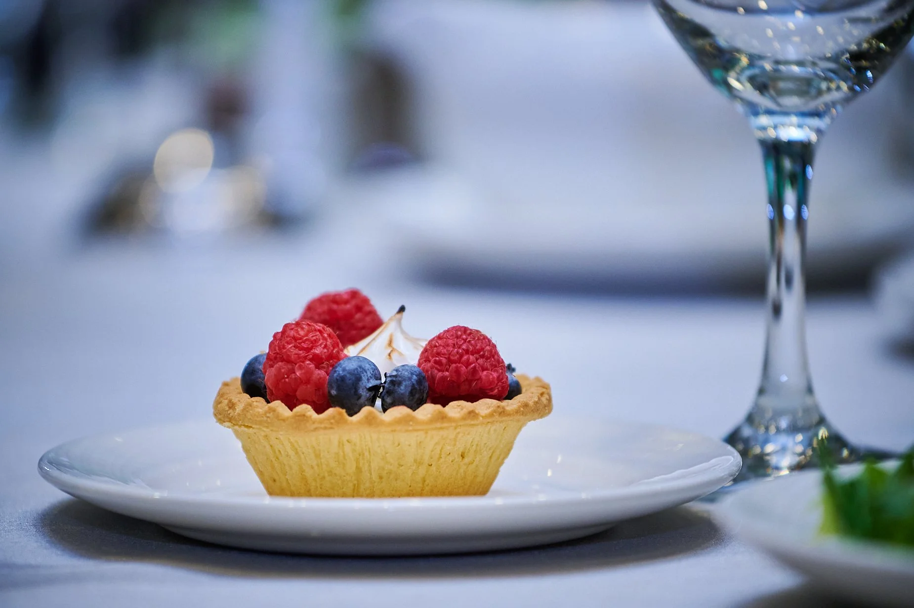 Fruit tart with raspberries and blueberries on a white plate, next to a wine glass.