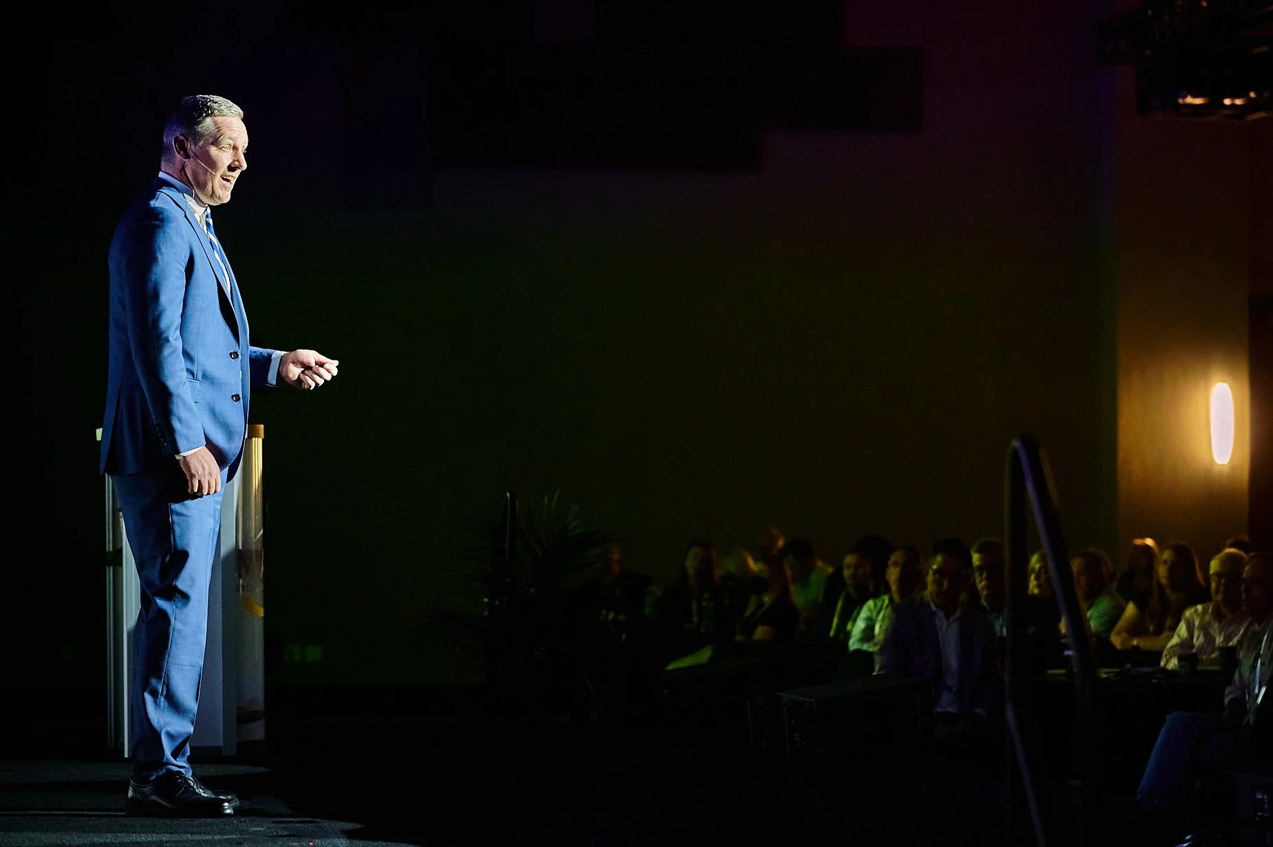 Man in blue suit speaking on stage at a conference with an audience in the background.