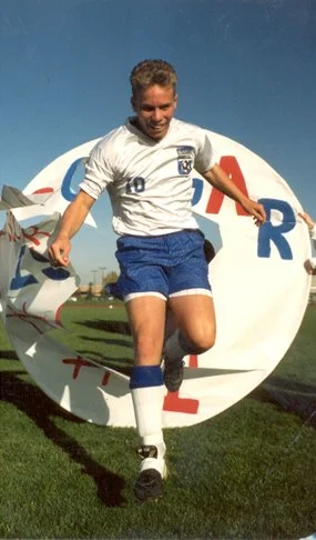 A soccer player in a white jersey and blue shorts is jumping in front of a circular sign with partial text, outdoors on a grass field.