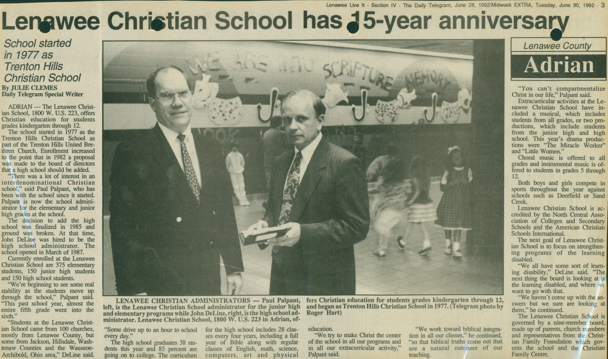 A newspaper article celebrating Lenawee Christian School's 15-year anniversary, featuring a black-and-white photo of two men in suits standing in a school hallway decorated with Christmas-themed artwork, with children in school uniforms in the background.