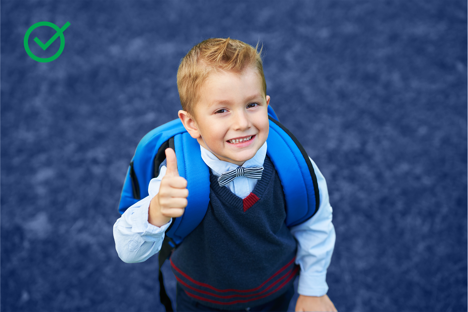 Smiling young boy wearing a school uniform, bow tie, and backpack, giving a thumbs-up gesture on a blue surface.