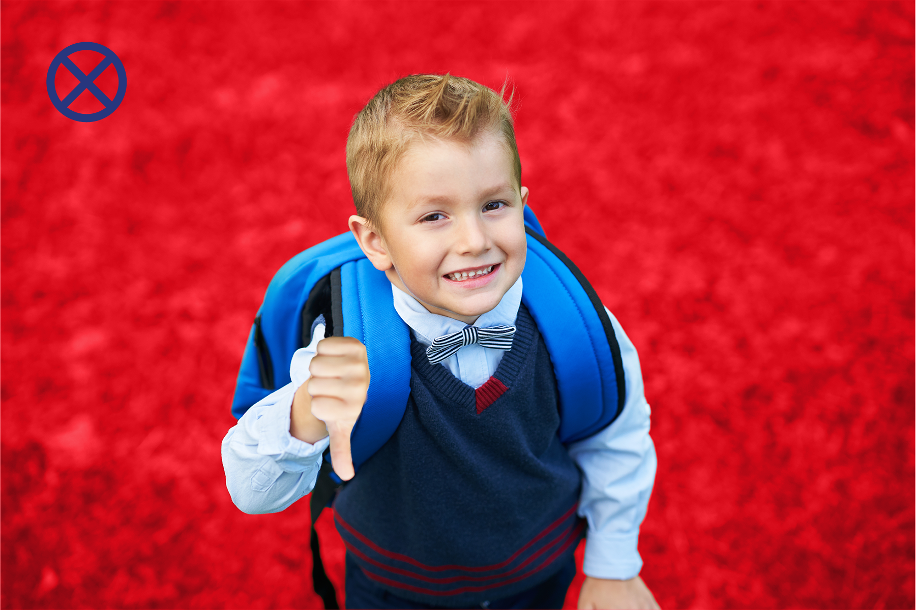 Young boy in school uniform giving thumbs down, wearing a blue backpack, smiling, on red sports field.