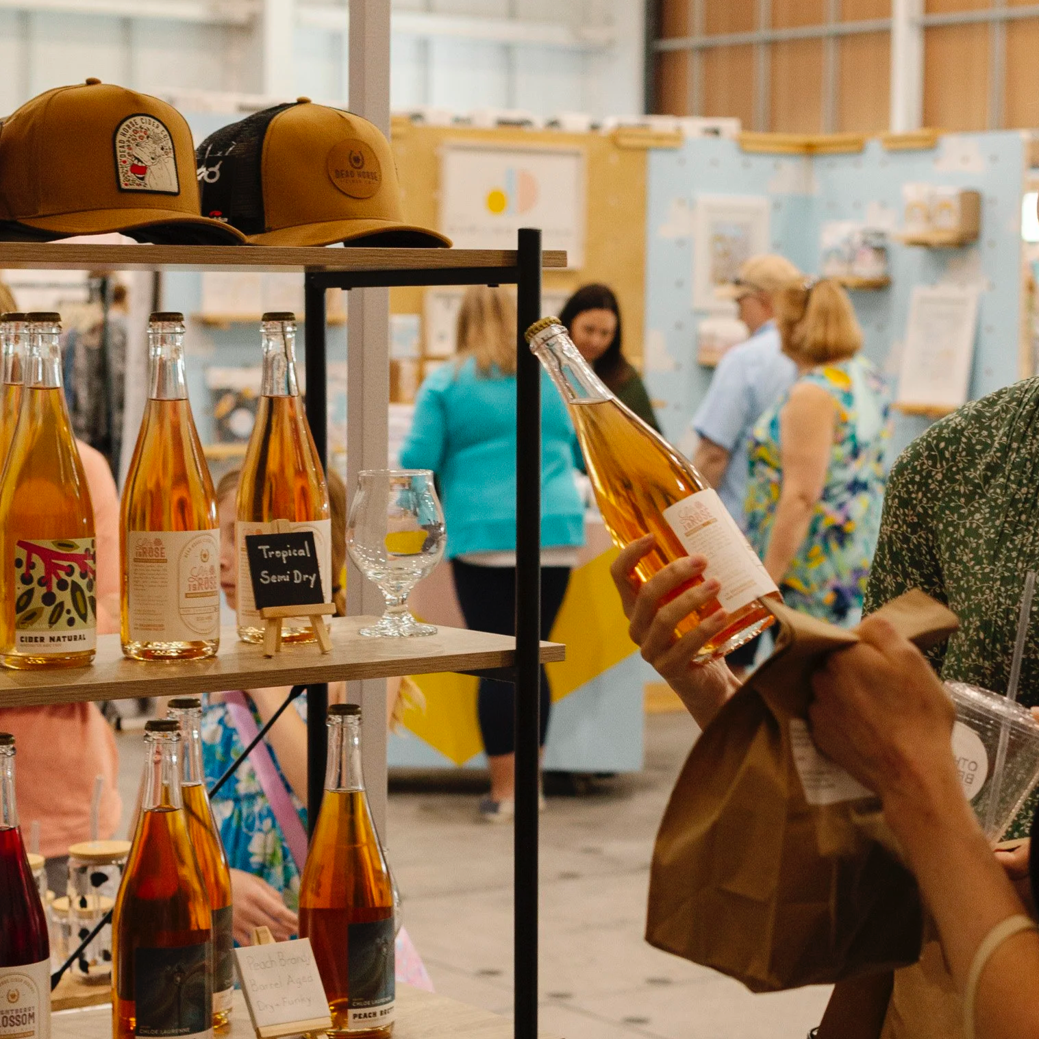 Person shopping for bottled beverages at an indoor market or store, with shelves displaying various bottles, some with labels and a small chalkboard sign reading 'Tropical Semi Dry'.