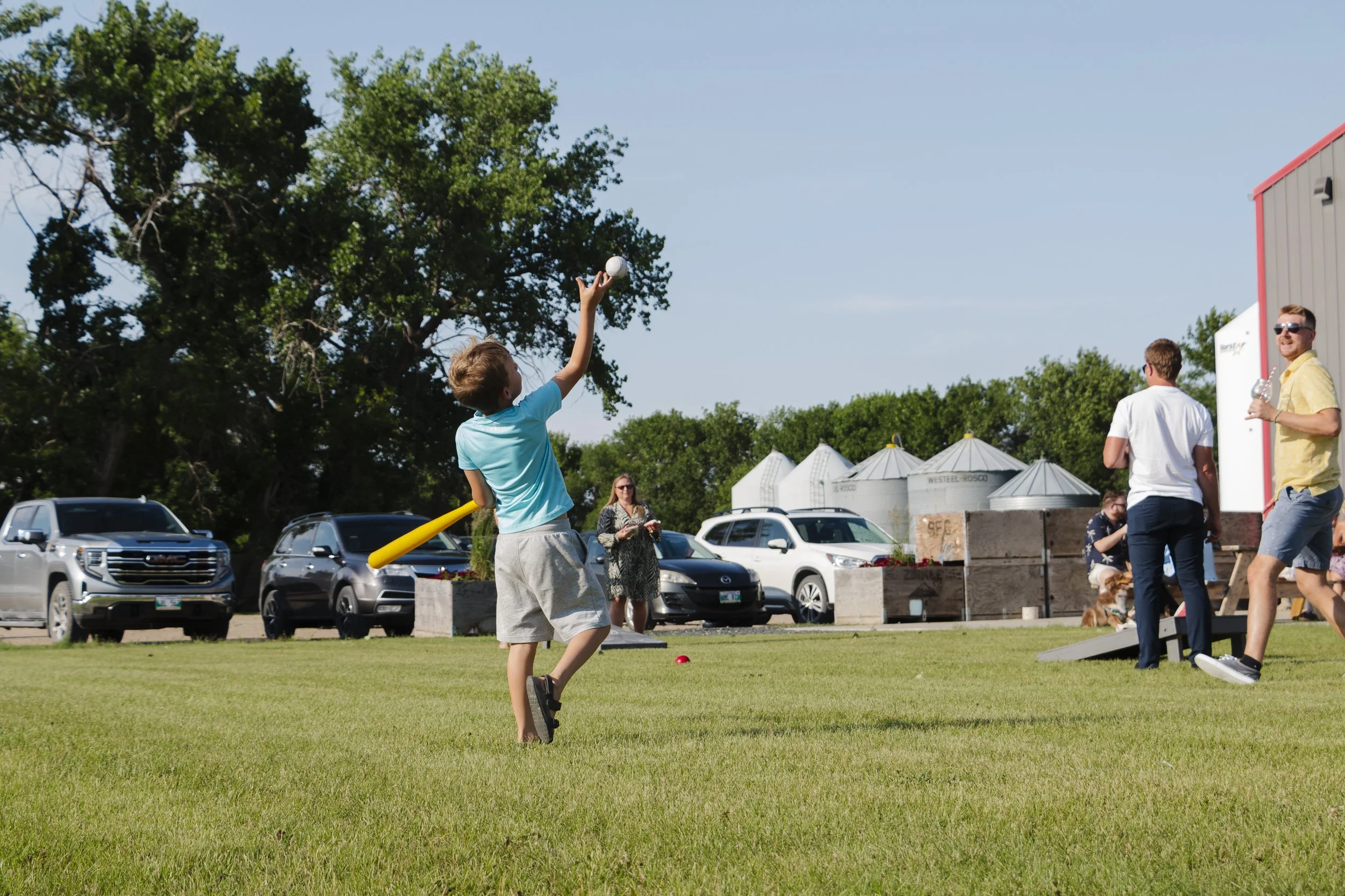Interior of a rustic-style restaurant with dark wooden tables and gray upholstered chairs. Potted plants and wooden barrels decorate the space, with a wooden wall and string lights in the background.