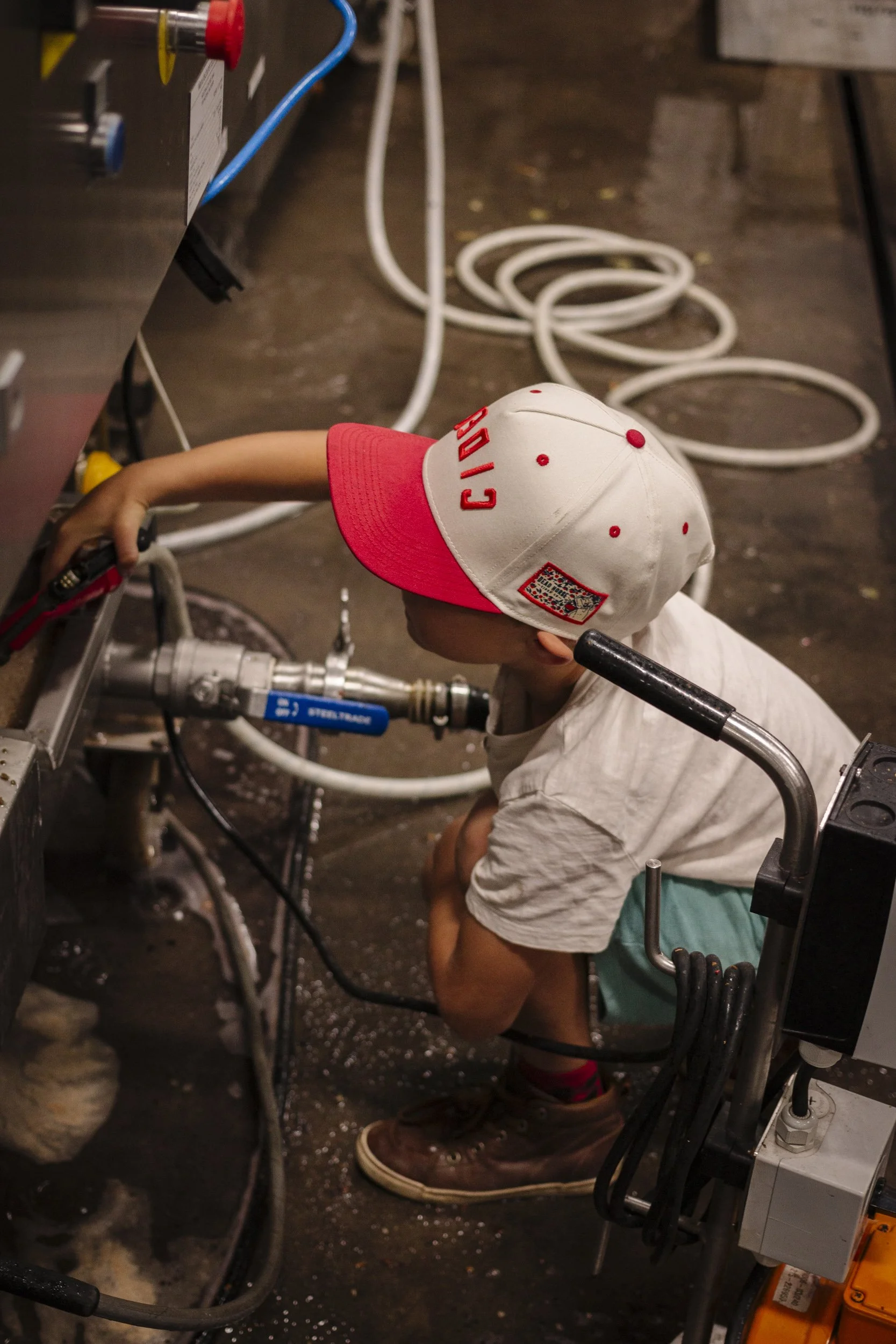 A young boy wearing a white and red cap and a white t-shirt, crouching down and working with machinery in an industrial setting, with various pipes and wires surrounding him.