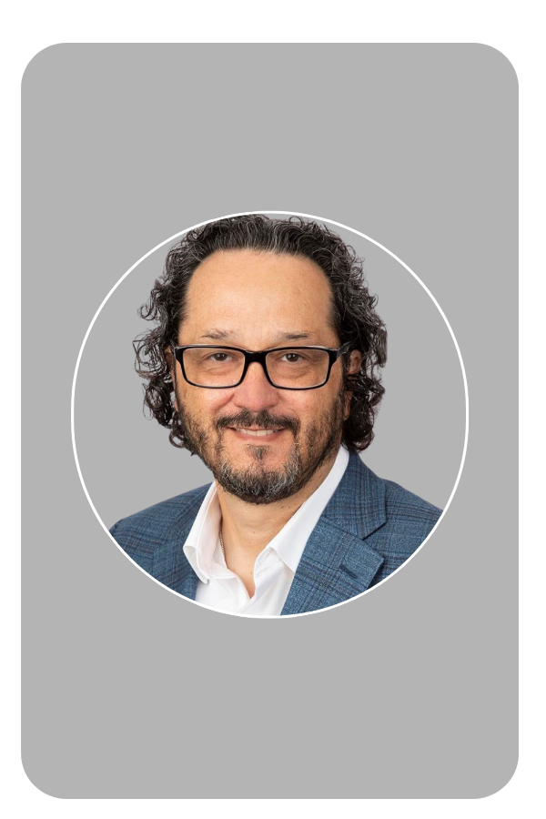 Man with glasses smiling, wearing a suit and shirt, dark background.
