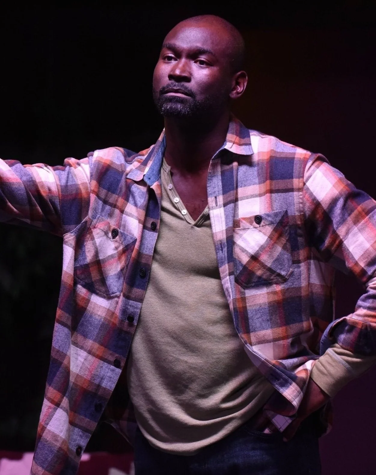 A man with a bald head and a beard, wearing a plaid shirt over a tan t-shirt, standing with one hand on his hip and the other extended, against a dark background.