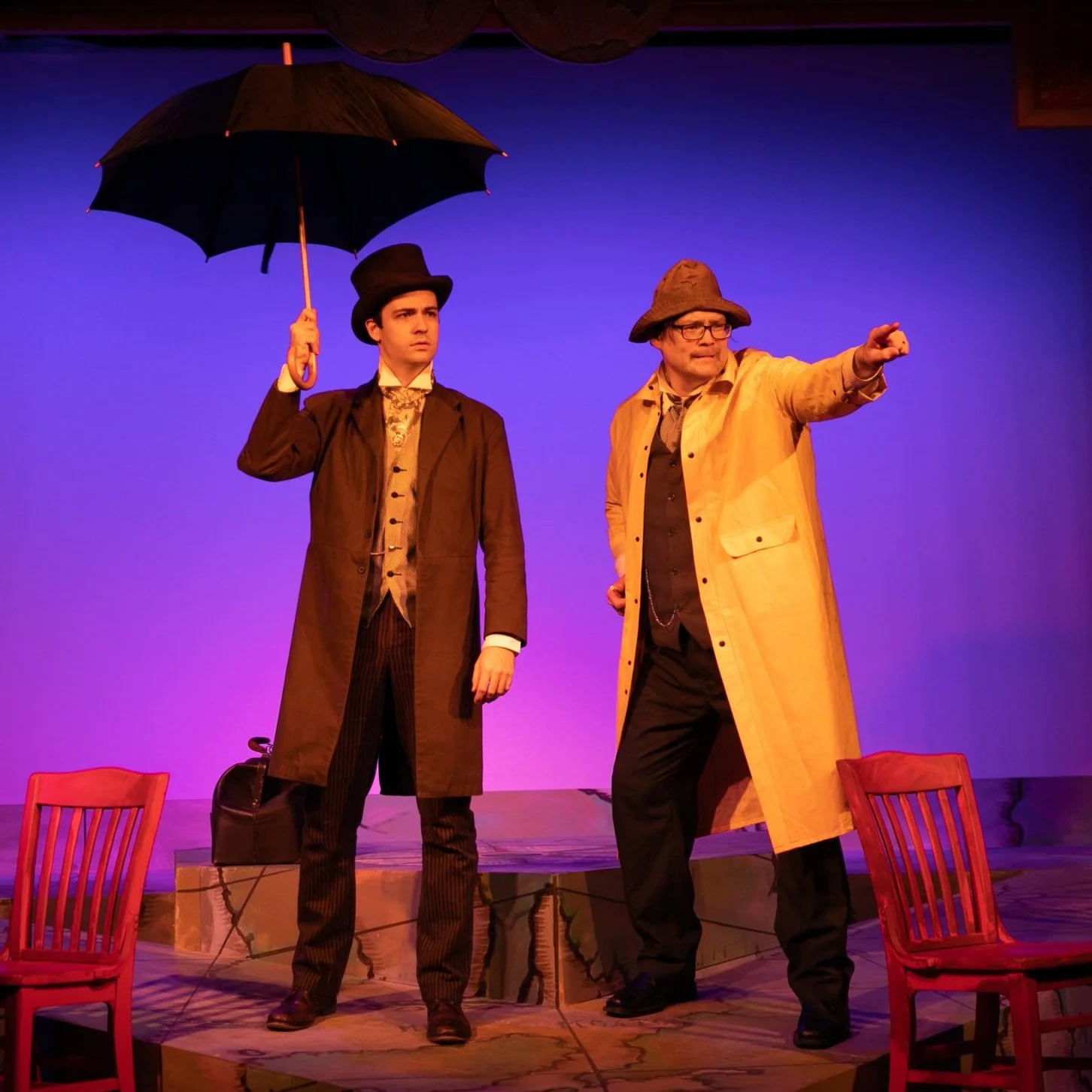 Two men in theatrical costumes on stage, one holding an umbrella and the other pointing, with chairs in the background and theatrical lighting.