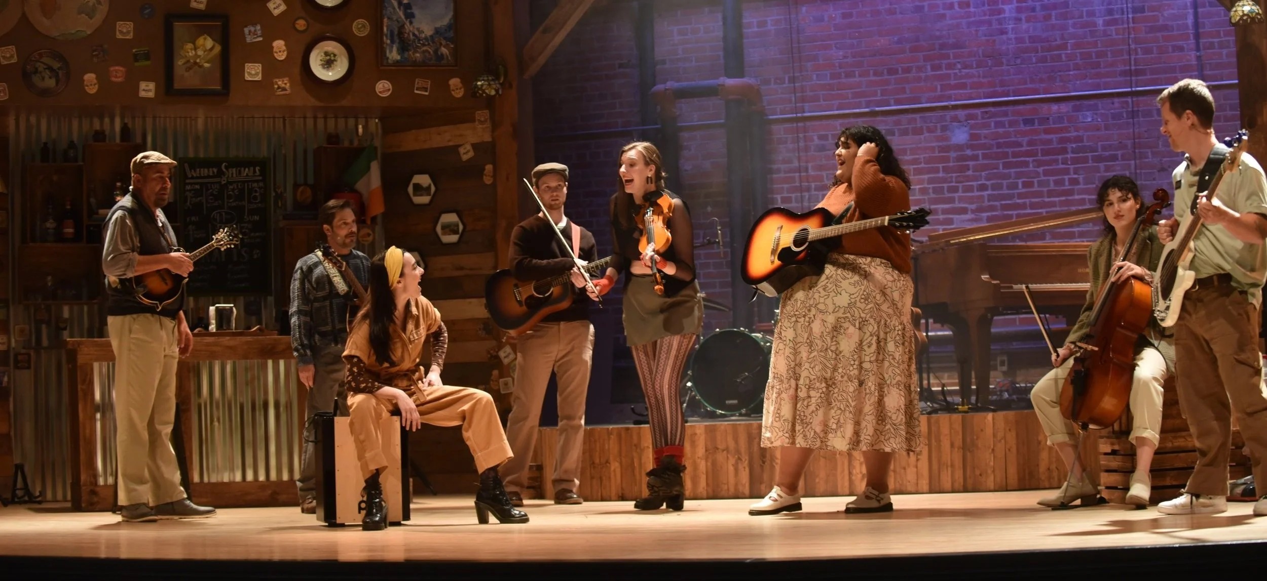 A group of musicians performing on stage in a rustic setting with brick walls, wooden accents, and wall decorations including an Irish flag. The group includes people playing guitars, a cello, and a keyboard, with one person sitting on a cajón drum. The scene depicts a lively musical session.