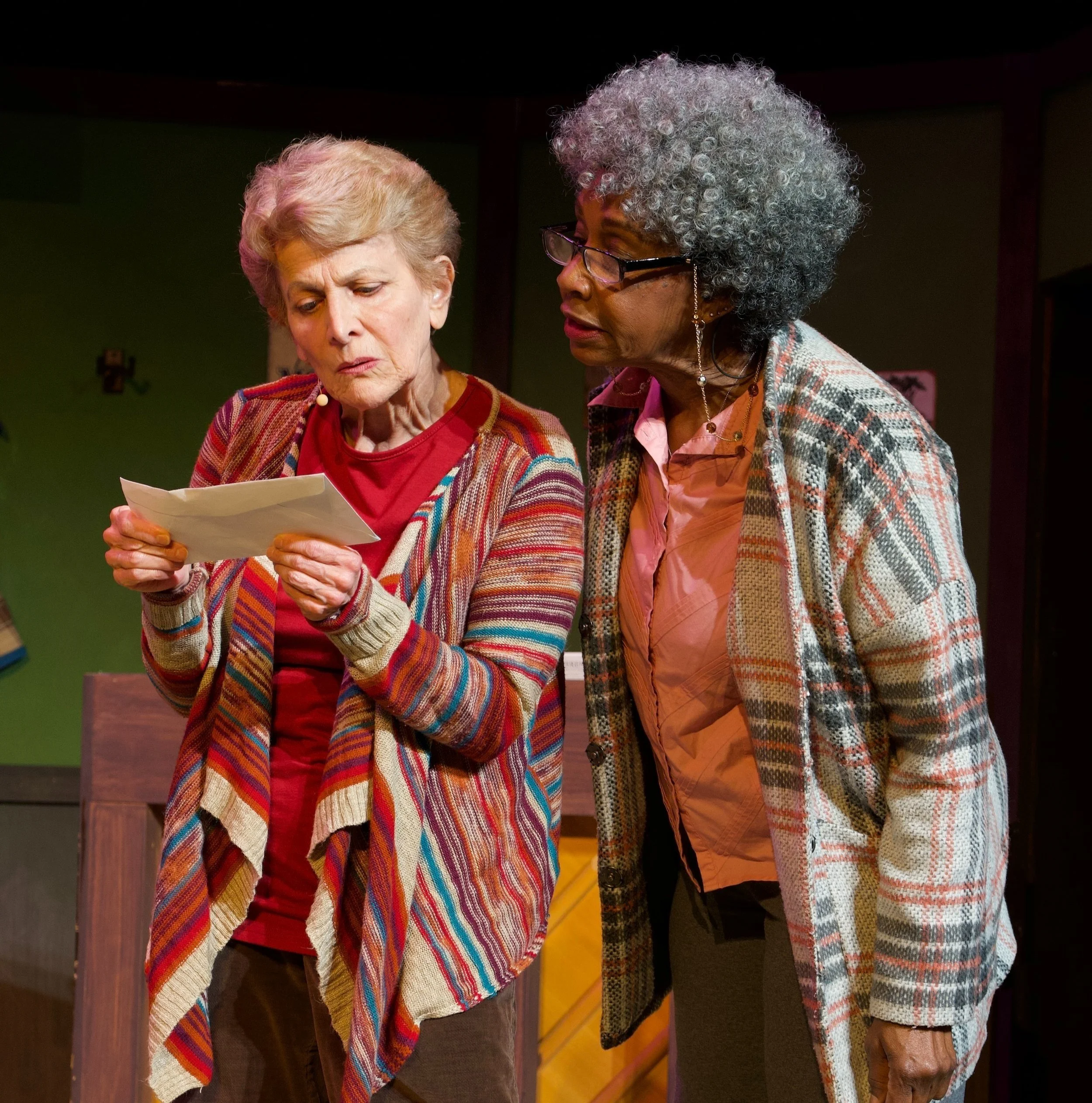 Two elderly women with gray and white curly hair, one with a beige and red plaid jacket and the other with a multicolored cardigan, stand close together looking at a piece of paper, indoors with a green wall and wooden furniture.