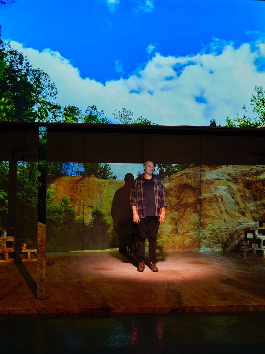 A man standing on a stage with a nature-themed backdrop, including trees, rocks, and a blue sky with clouds, with lighting creating a shadow of the man on the backdrop.