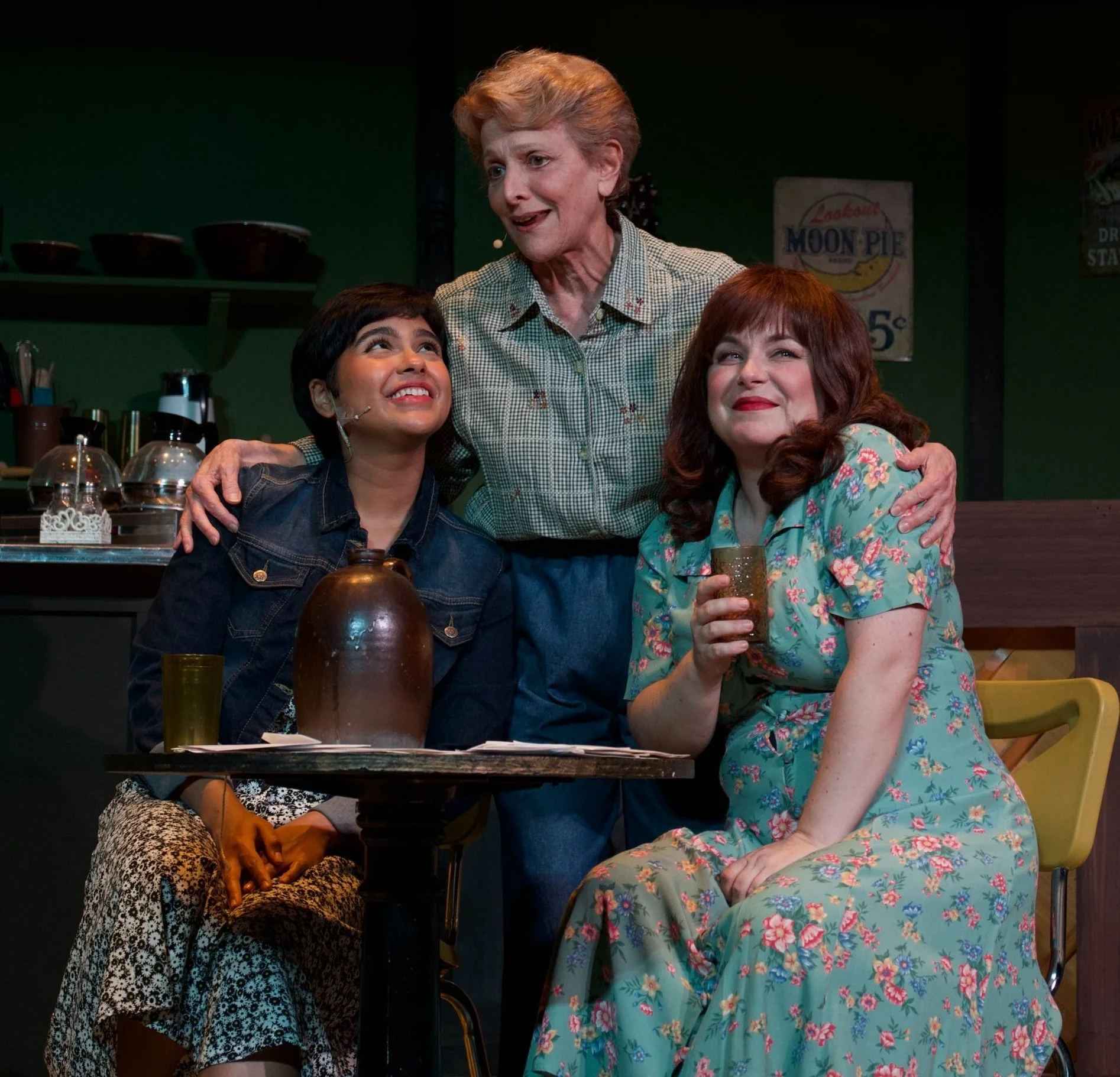 Three women on stage, one older woman standing and two younger women sitting, smiling and engaging, in a cozy, vintage-style diner setting with green walls and retro decor.