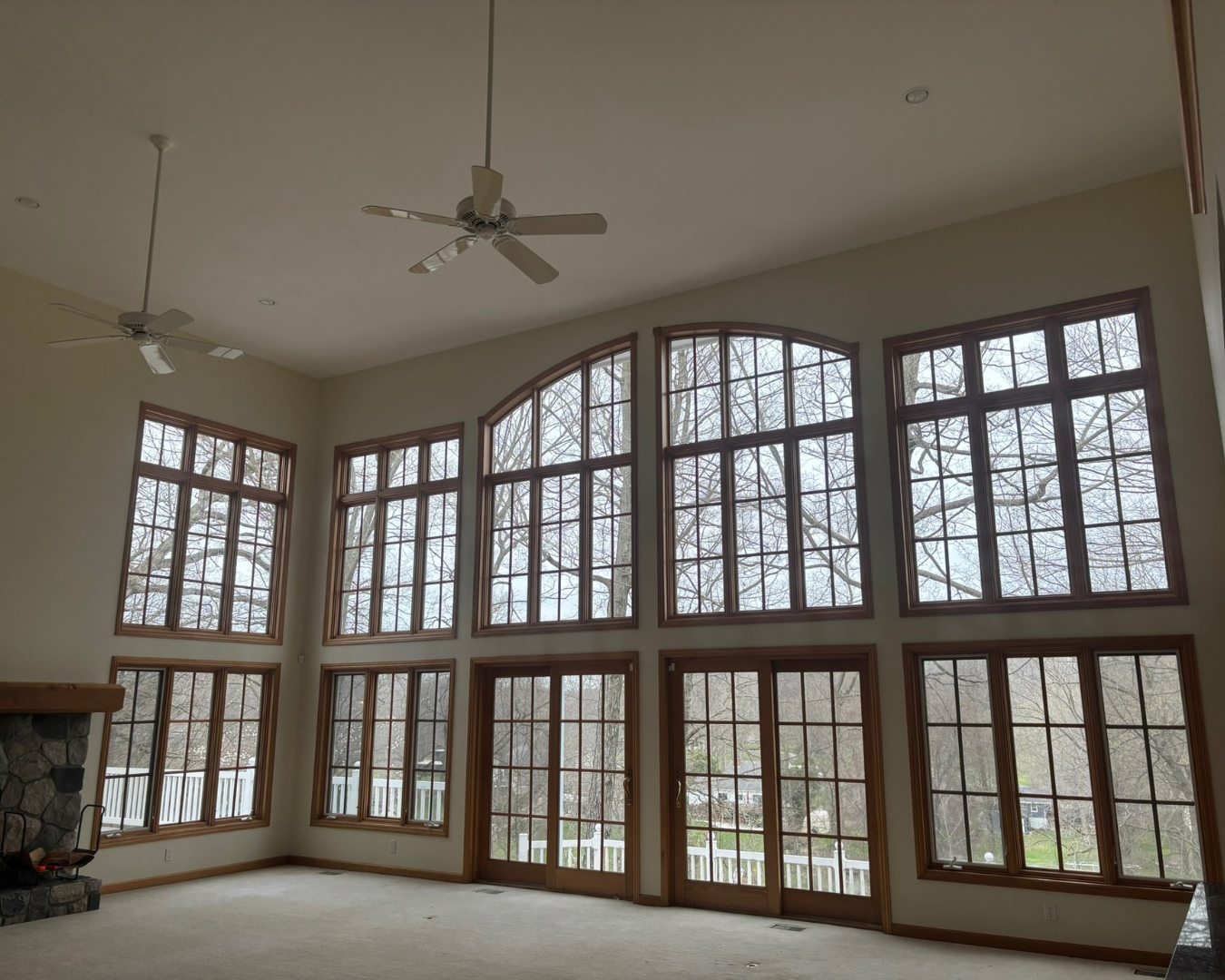 Large living room with tall, multi-pane windows and a glass door, showing trees outside, with white walls, ceiling fans, and a stone fireplace.