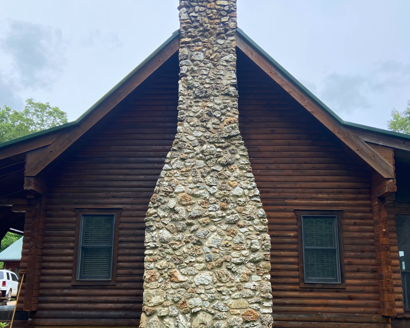 Close-up of a lodge-style house with a prominent stone chimney running through the center, wooden logs making up the walls, and two windows with shutters. Part of a white vehicle is visible on the left side.