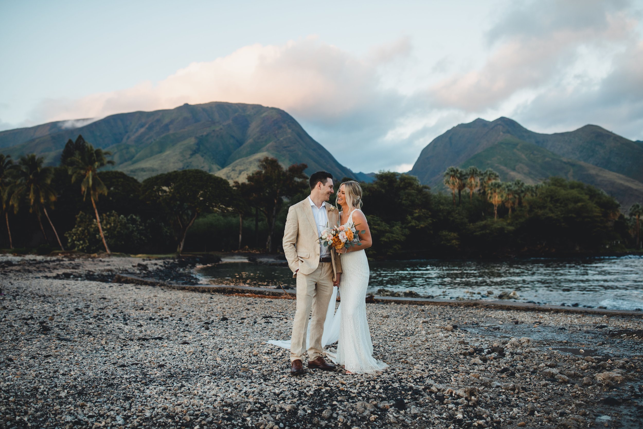A bride and groom on a lava rock beach, with a majestic West Maui Mountain range backdrop.