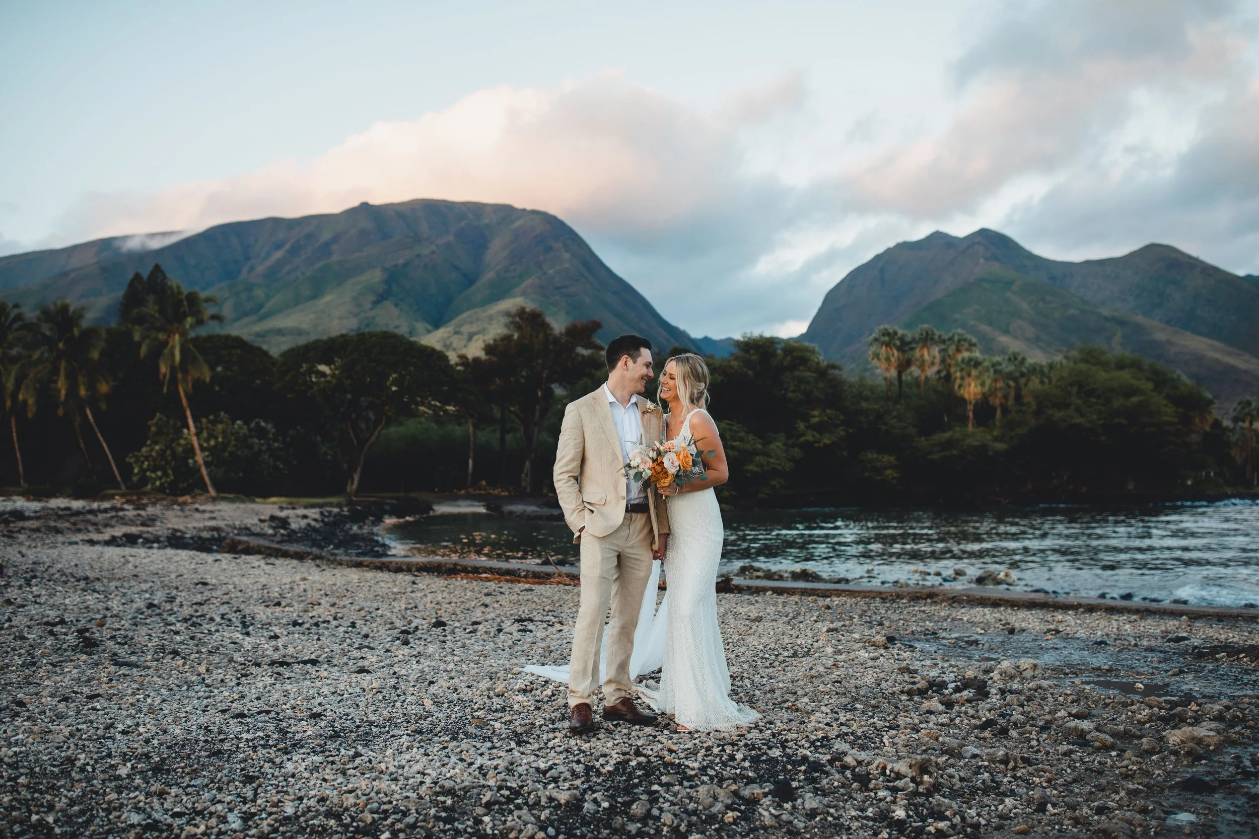 A bride and groom on a lava rock beach, with a majestic West Maui Mountain range backdrop.