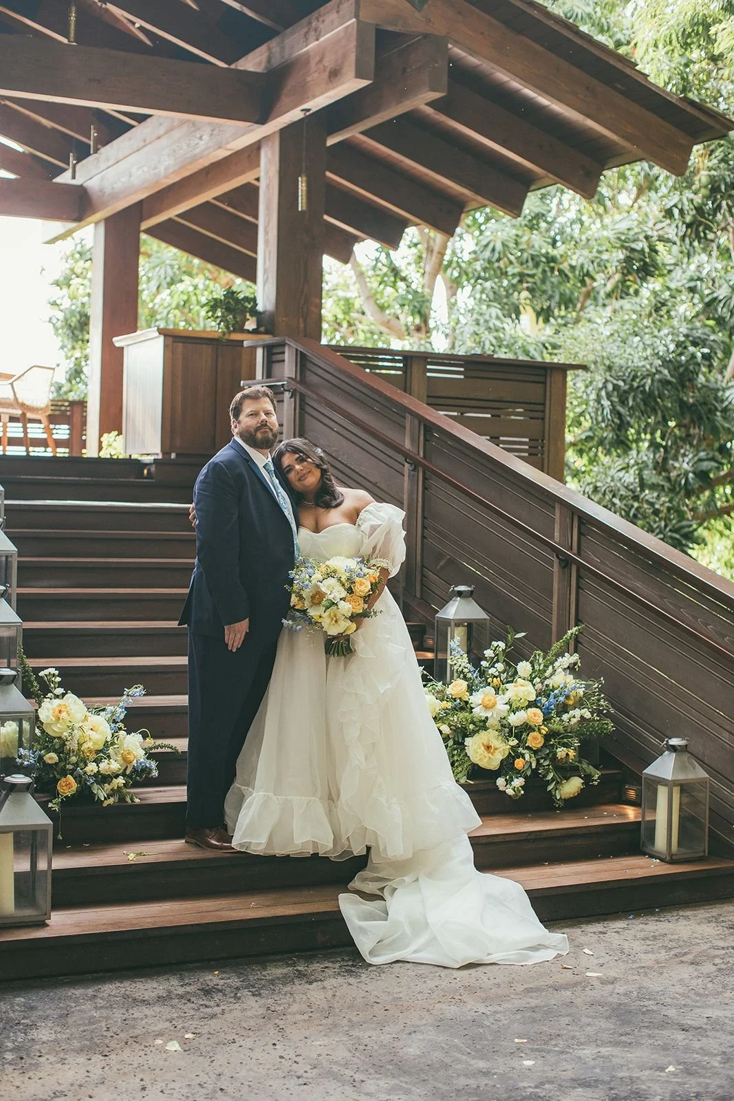 A bride and groom standing at an outdoor wooden staircase between large wedding flower installations