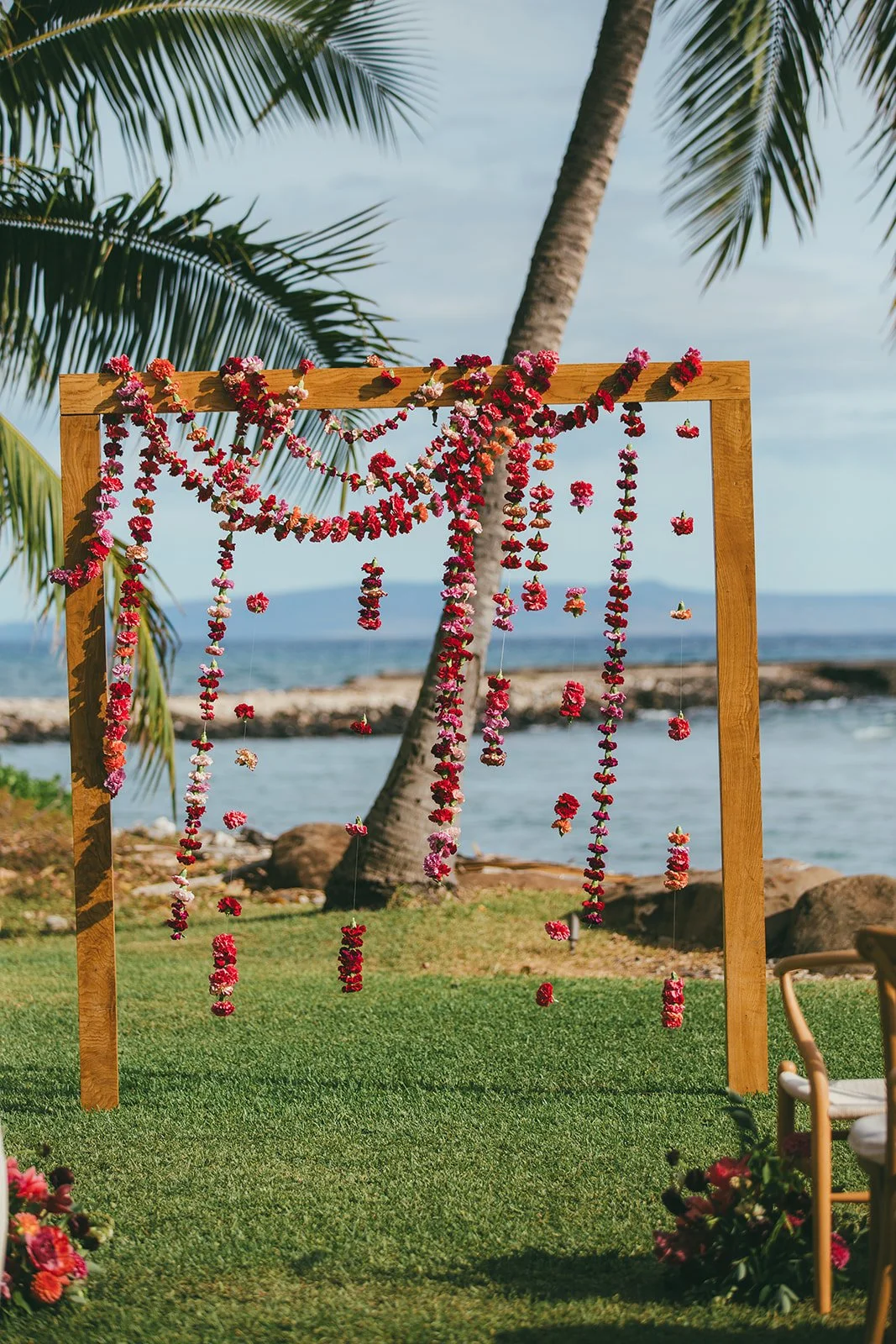 A floral wedding ceremony on the beach, with an angular wooden arch draped with fuchsia carnations and jewel-tone floral aisle markers.
