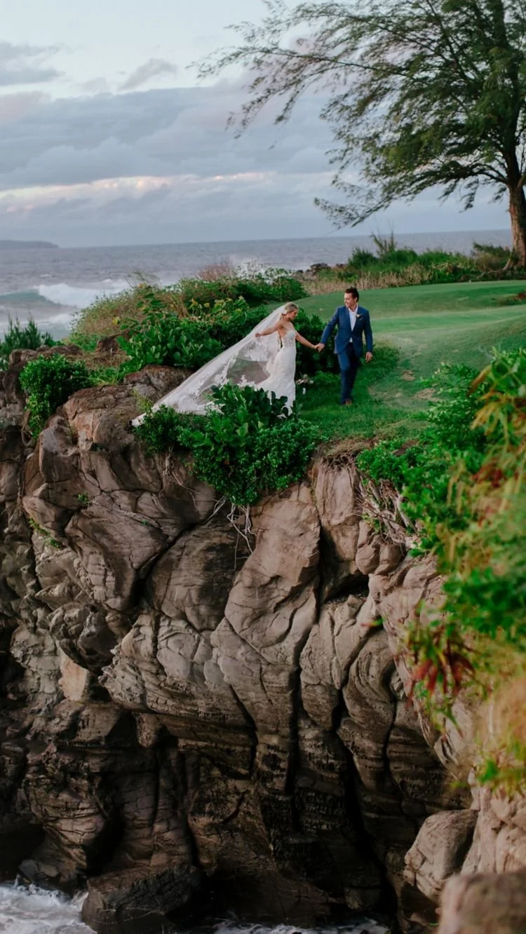 A bride and groom at a grassy cliffside over the pale blue ocean.
