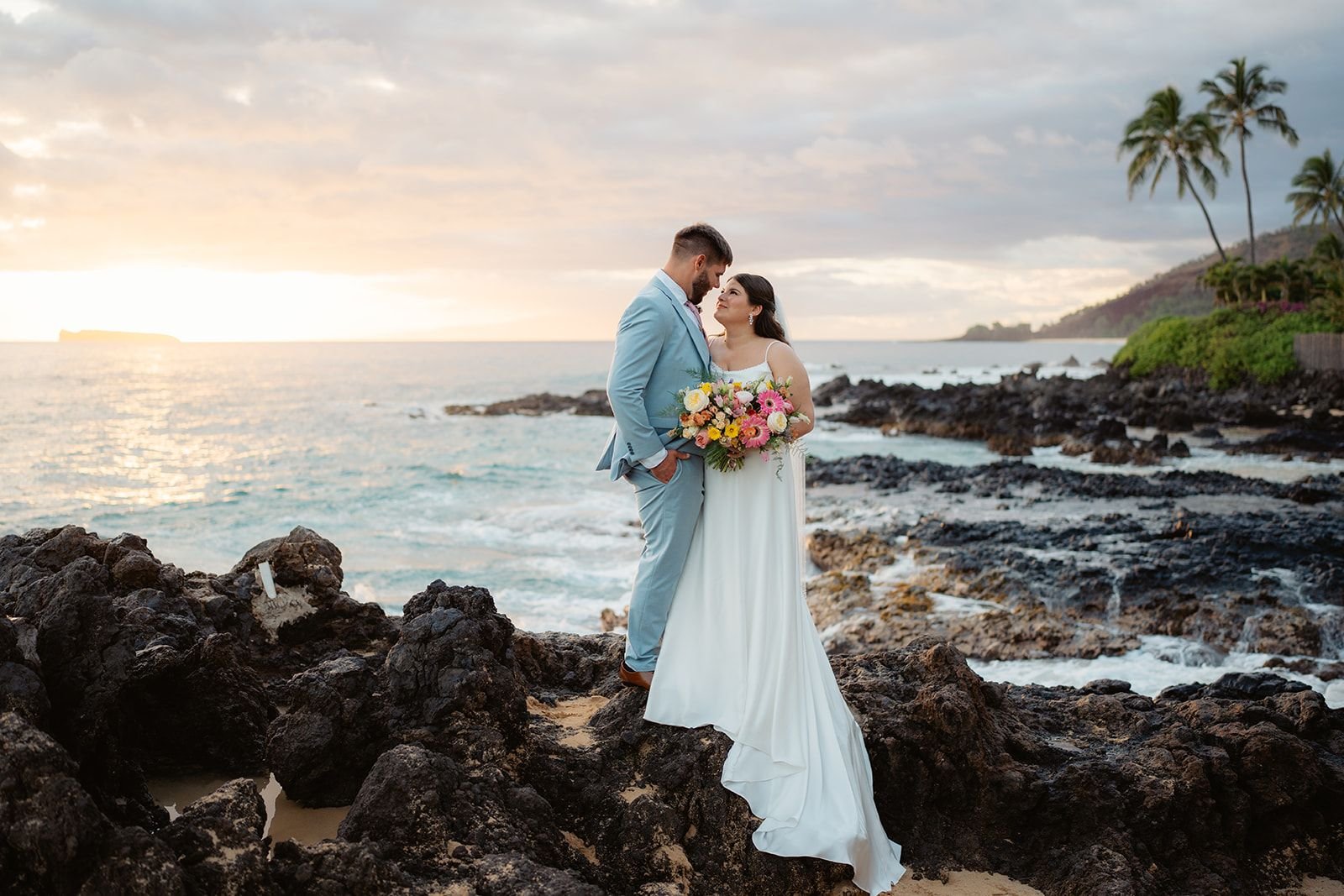 A bride and groom standing on lava rock overlooking a Maui ocean cove. The bride carries a bright spring bouquet.