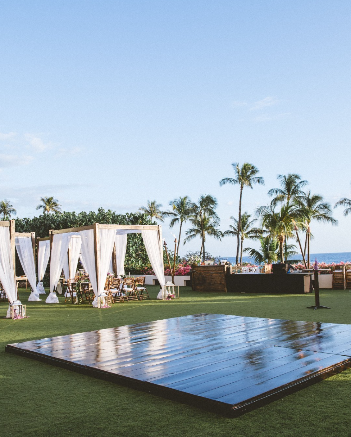 An outdoor wedding reception on the grass with a dance floor in the foreground and canopied tables and palm trees in the background.