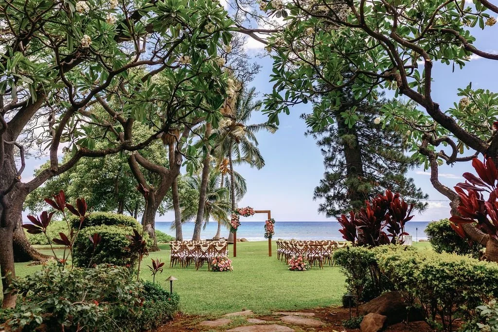A vibrant tropical oceanside wedding on the grass surrounded by ti leaves, plumeria, pine, and palm trees.